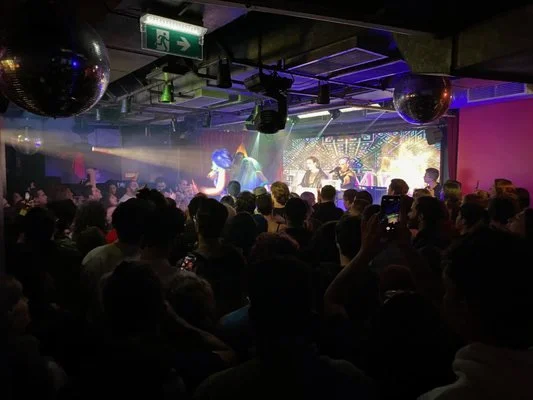 Crowd watching a live band perform on stage at a nightclub with colorful lighting and a disco ball overhead.