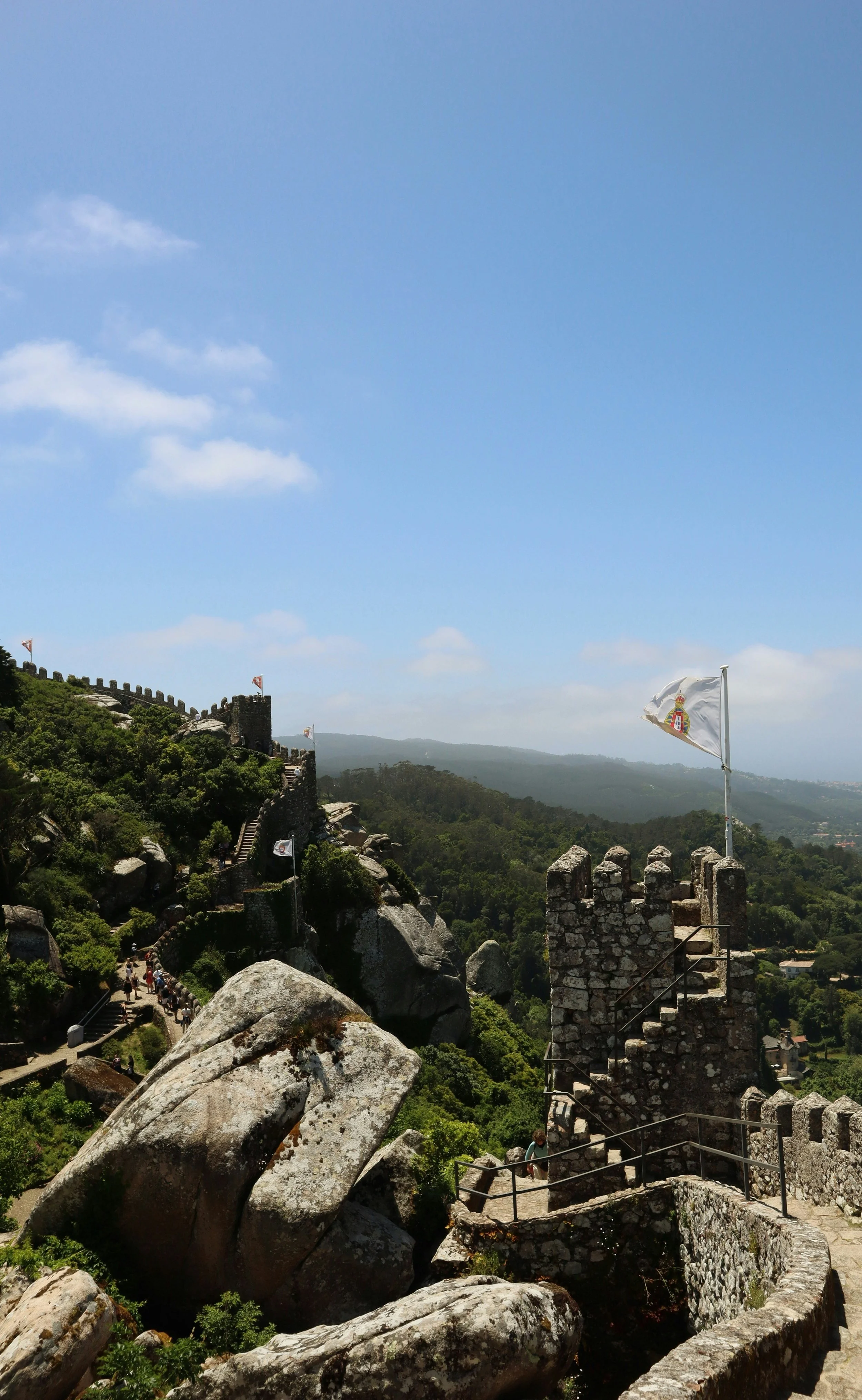 A scenic view of a historic fortress with stone walls and flags flying, surrounded by green hills and rocks, under a partly cloudy blue sky.