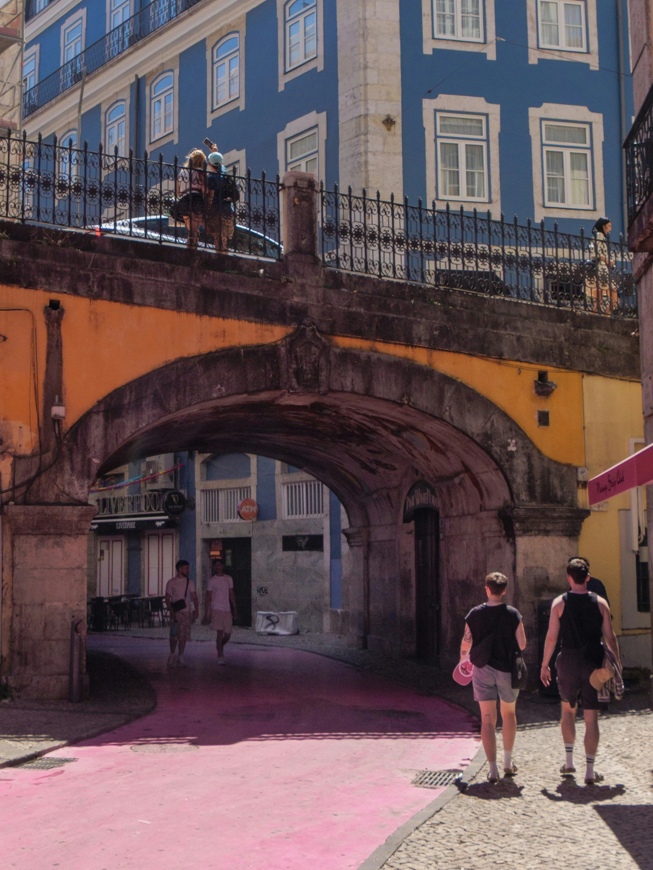 People walking on a street with a colorful building in the background, some standing on a bridge or archway, with bright sunlight and shadows casting on the ground.