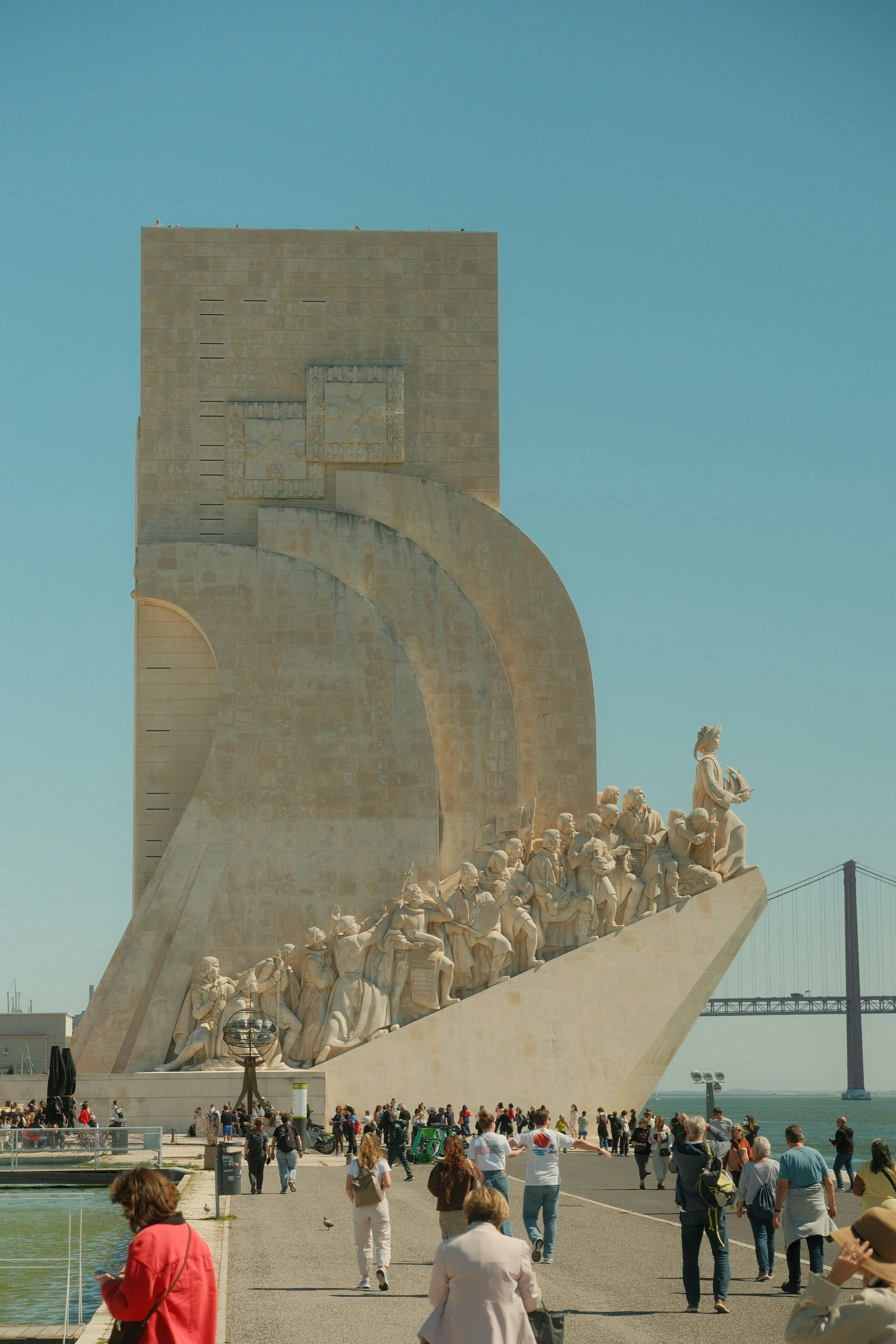 The Padrão dos Descobrimentos, a large stone monument with a sculpture of explorers and historical figures in Lisbon, Portugal, with people walking around and the 25 de Abril Bridge in the background.