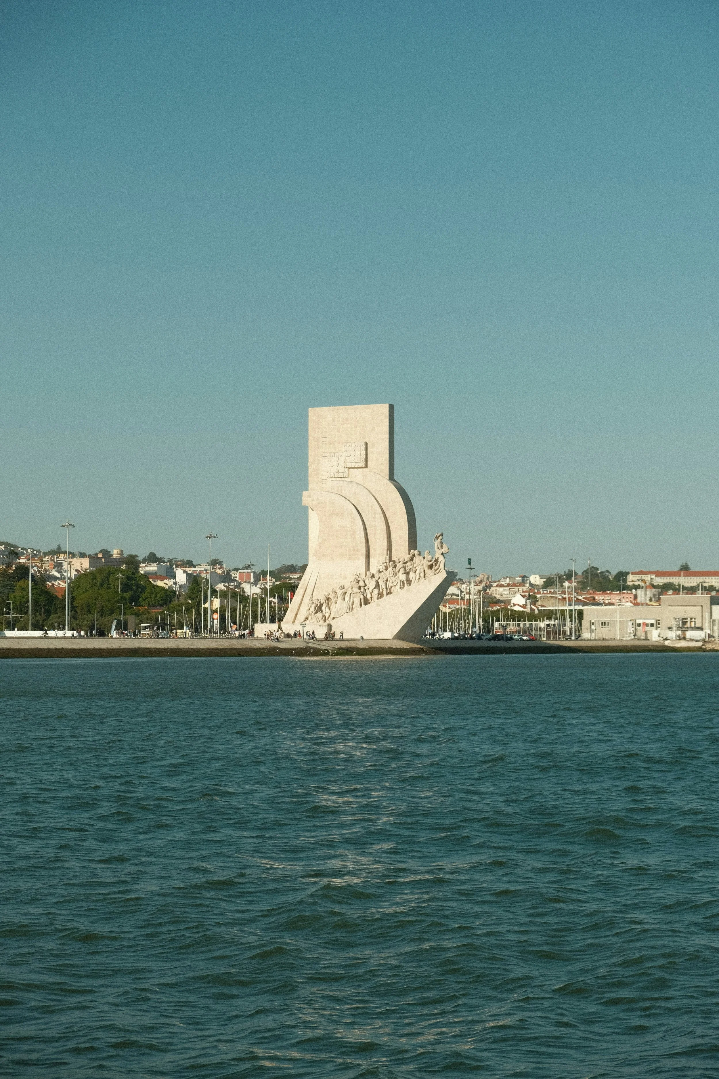 A large white monument with sculptures at its base, situated by the water with a cityscape and hills in the background, under a clear blue sky.