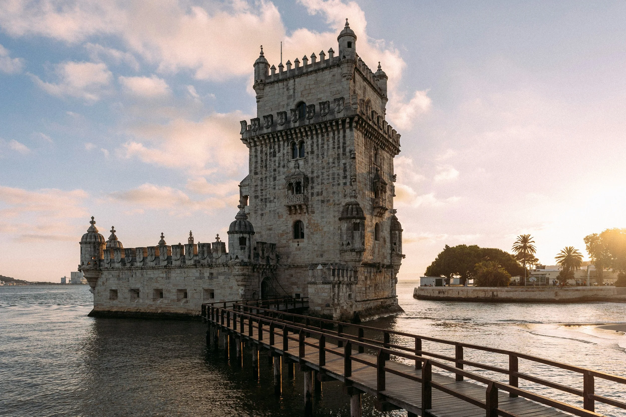 Historic stone fortress with towers located by the water, with a wooden walkway leading to it, palm trees and a partly cloudy sky with sunset lighting.