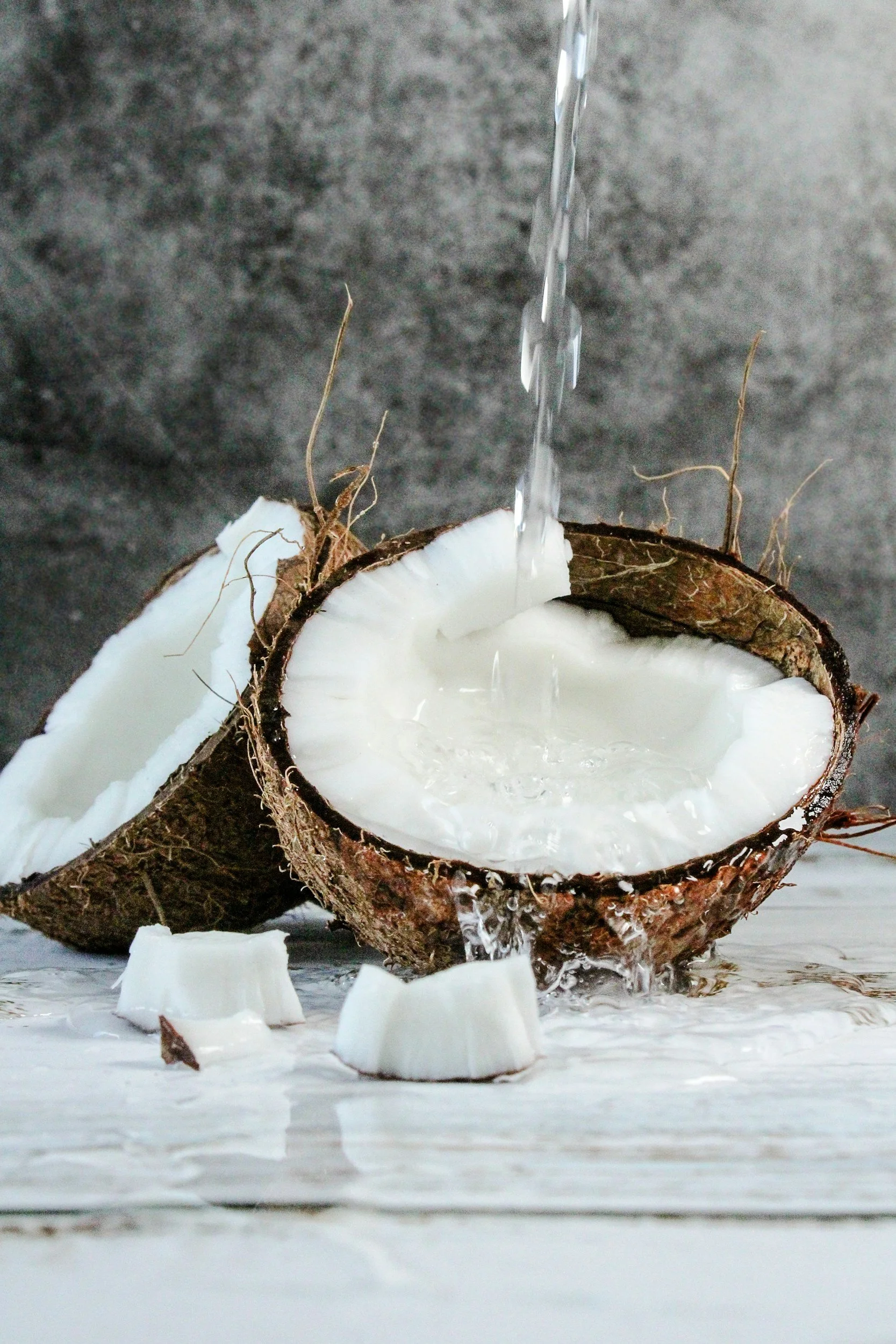 Una cáscara de coco cortada por la mitad con agua de coco vertiéndose en su interior, con pedazos de coco y cáscara en la superficie.