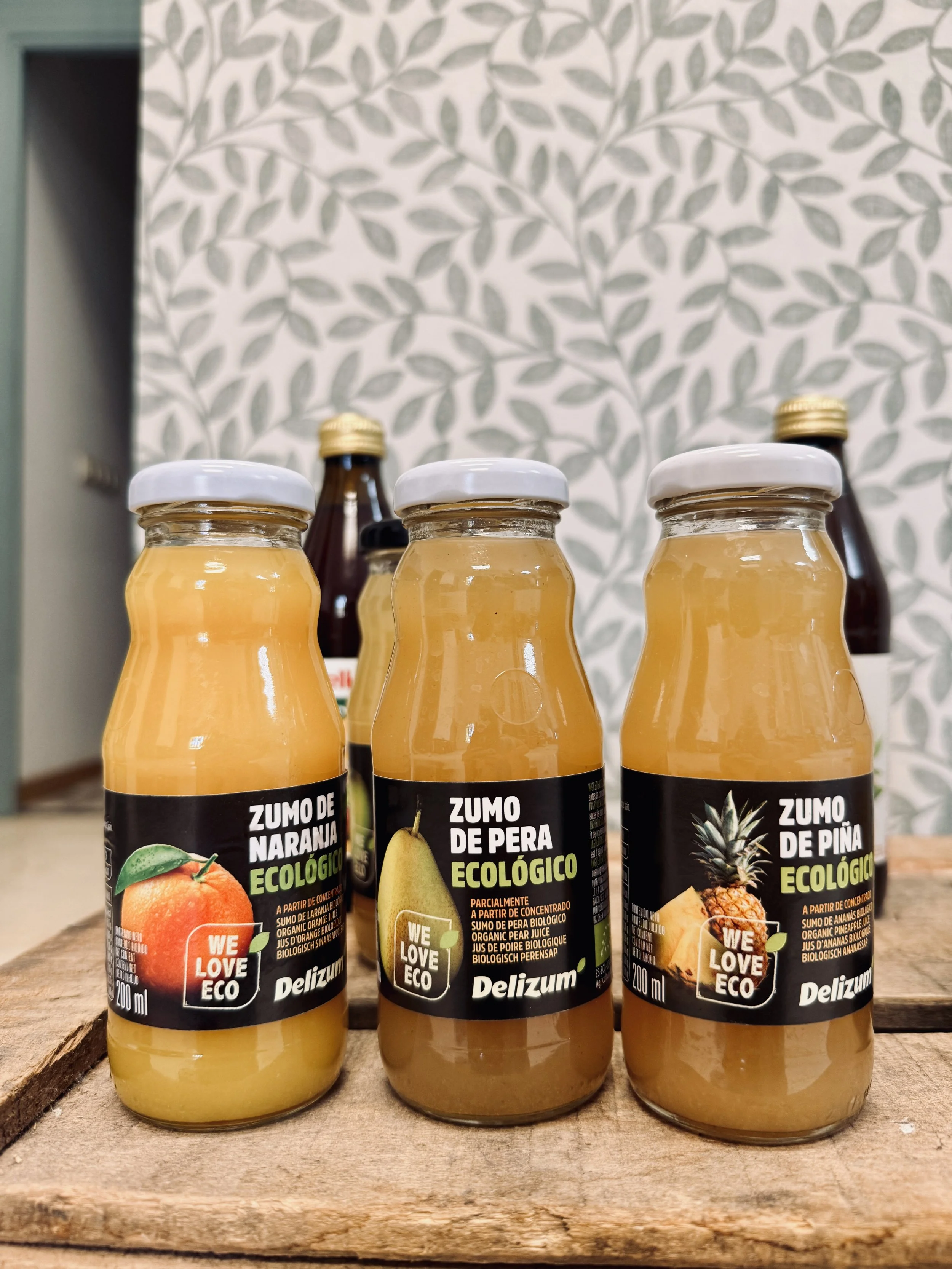 Frascos de jugo ecológico de diferentes frutas: naranja, pera y piña, con etiquetas en español y logotipo 'WE LOVE ECO', sobre una tabla de madera con fondo de papel tapiz con diseño de hojas grises.