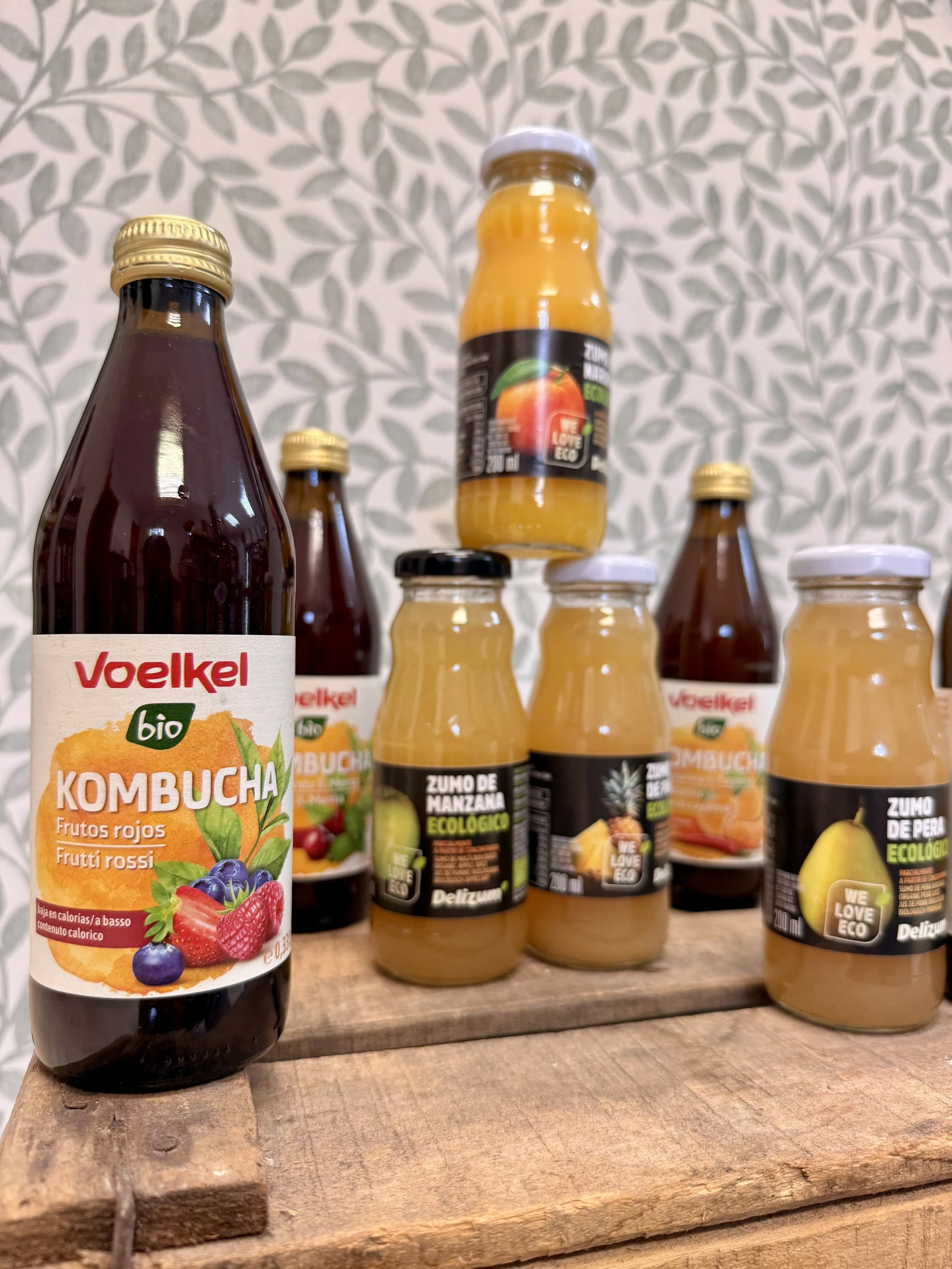 Botellas de jugo de frutas y kombucha en una estantería de madera con fondo de patrón de hojas en tonos blancos y gris.