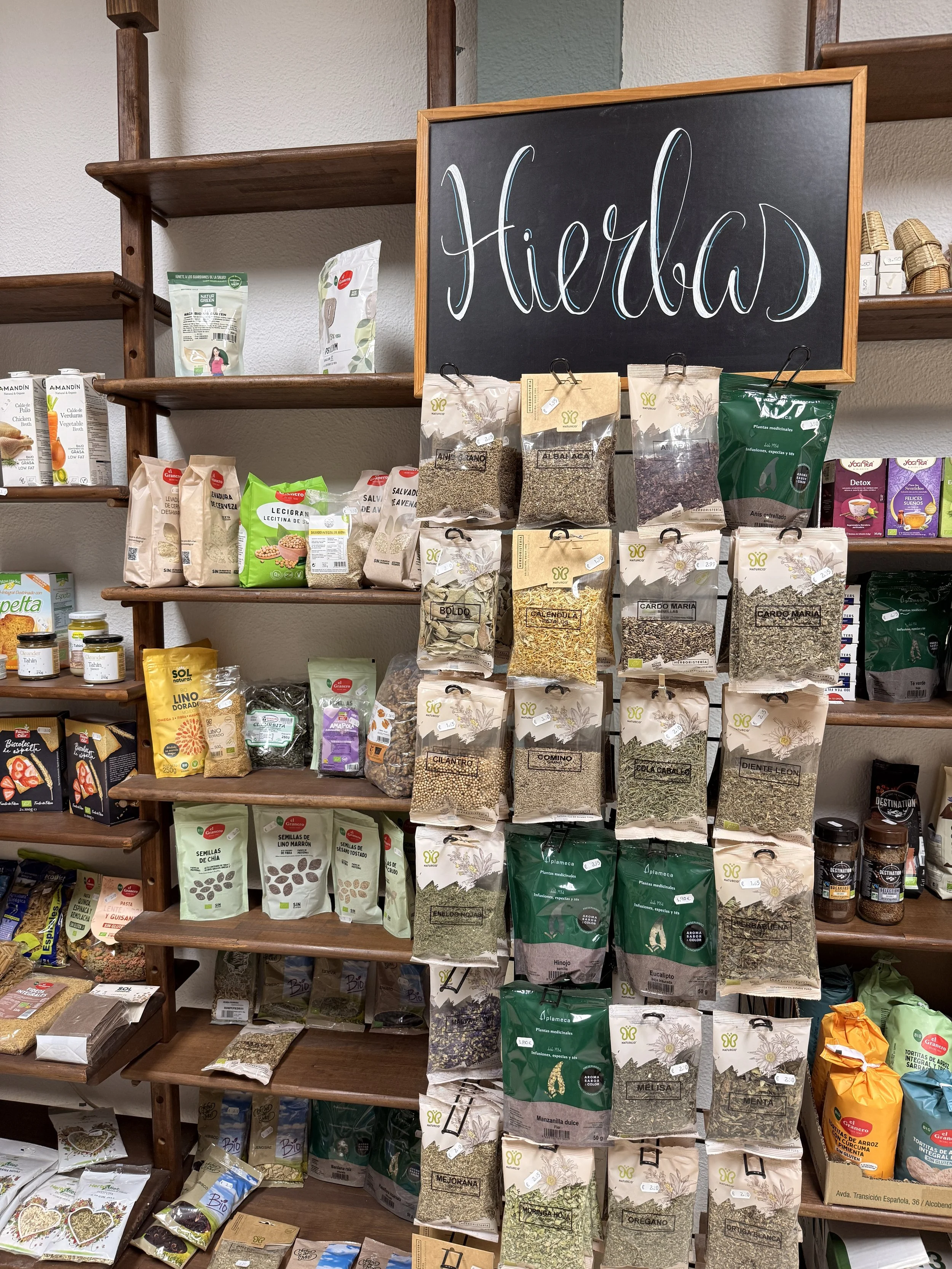 Estante de productos a granel y especias en una tienda natural, con un cartel que dice 'Hierbas' en letras tachadas, en español.
