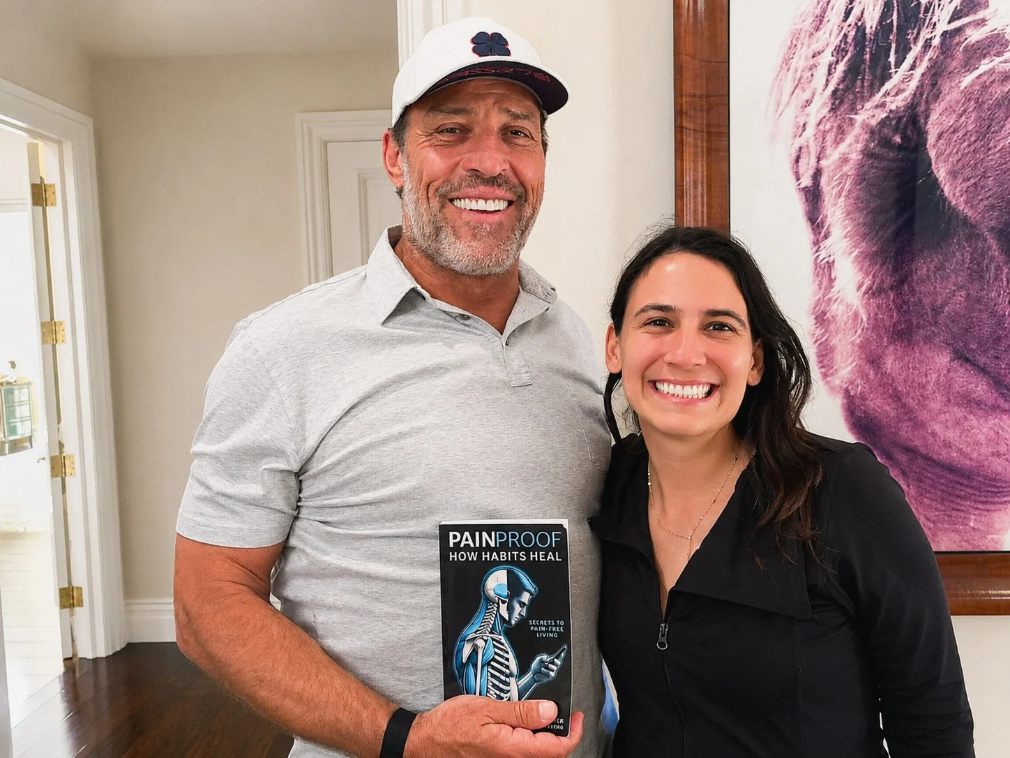 A smiling man and woman standing together indoors, holding a book titled 'Pain Proof' with a skull illustration, in front of a pink abstract painting.