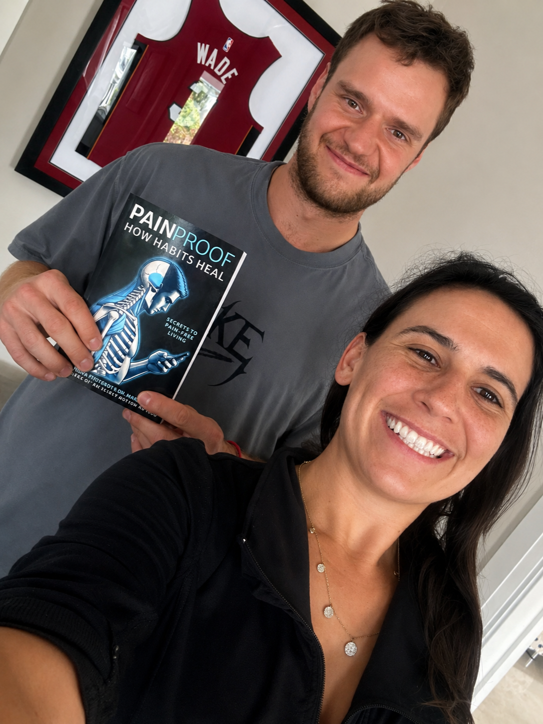 A smiling woman taking a selfie with a man holding a book titled 'Painproof: How Habits Heal'. The man is wearing a gray shirt and the woman is in a black zip-up jacket. In the background, a framed basketball jersey with the name 'Wade' is visible.
