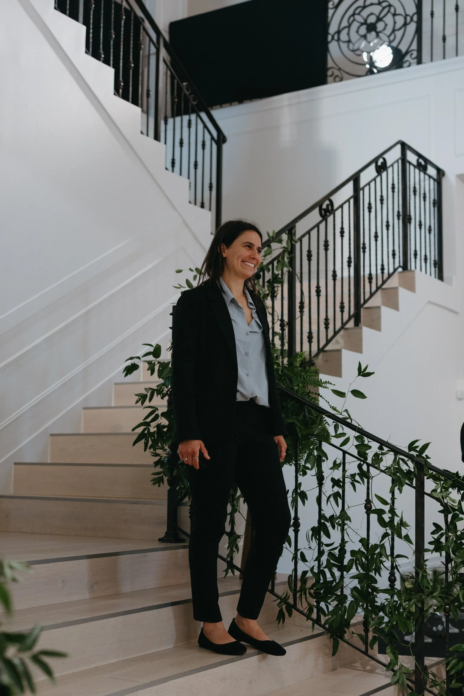 A woman with dark hair wearing a blazer, shirt, and black pants standing on a staircase with black railings and green plants nearby, in a well-lit indoor space.