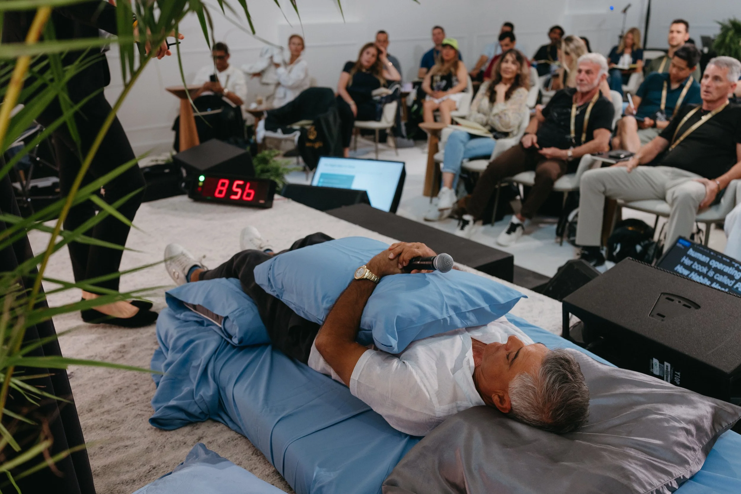 A man lying on a hospital bed giving a presentation with a microphone in his hand, surrounded by an audience in a conference room.