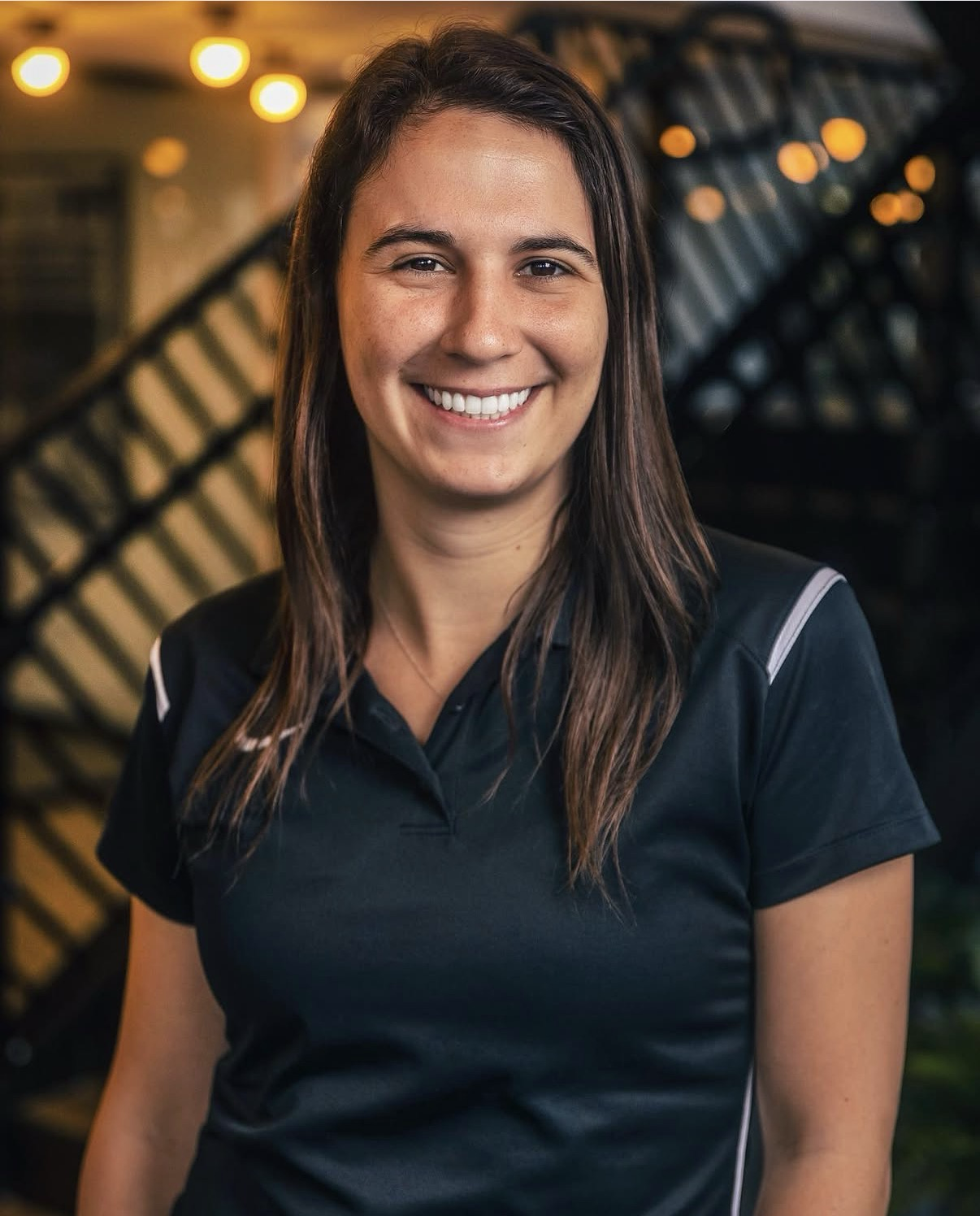 A smiling woman with brown hair wearing a black sports shirt standing in front of string lights and a building.