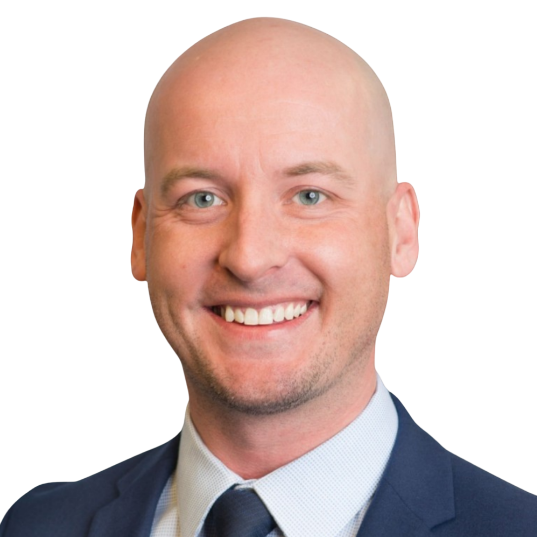 A smiling man with a bald head, light skin, blue eyes, dressed in a navy suit, white shirt, and light-colored tie.