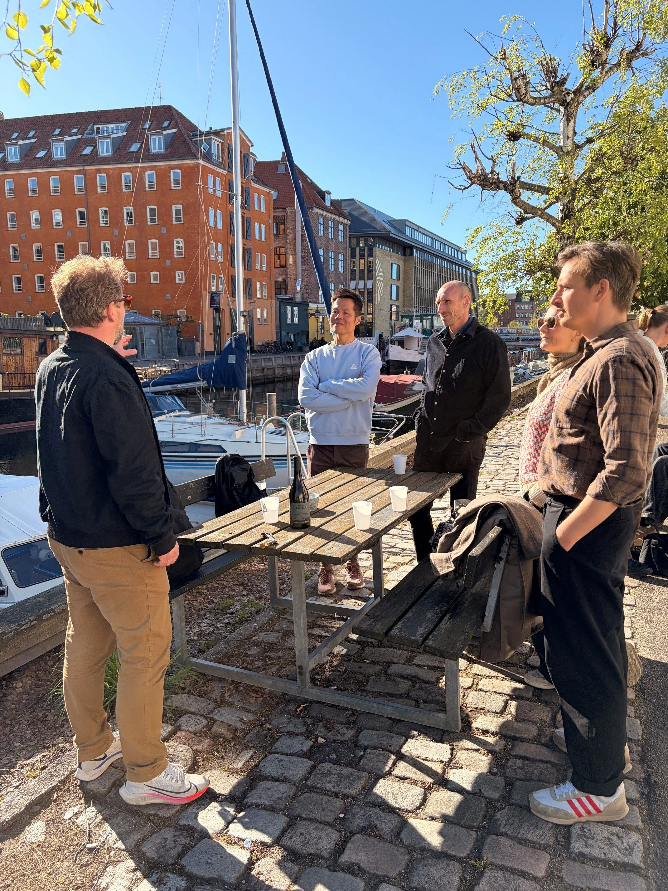 A group of five people gathered around a wooden table outdoors near a marina, with sailboats and colorful buildings in the background, on a sunny day.
