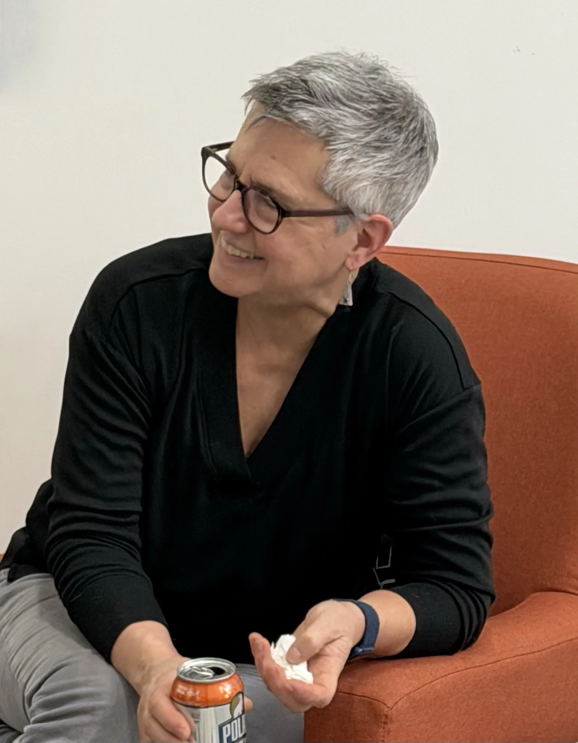 A smiling man with gray hair and glasses sits on an orange couch, holding a soda can in one hand and a napkin in the other.