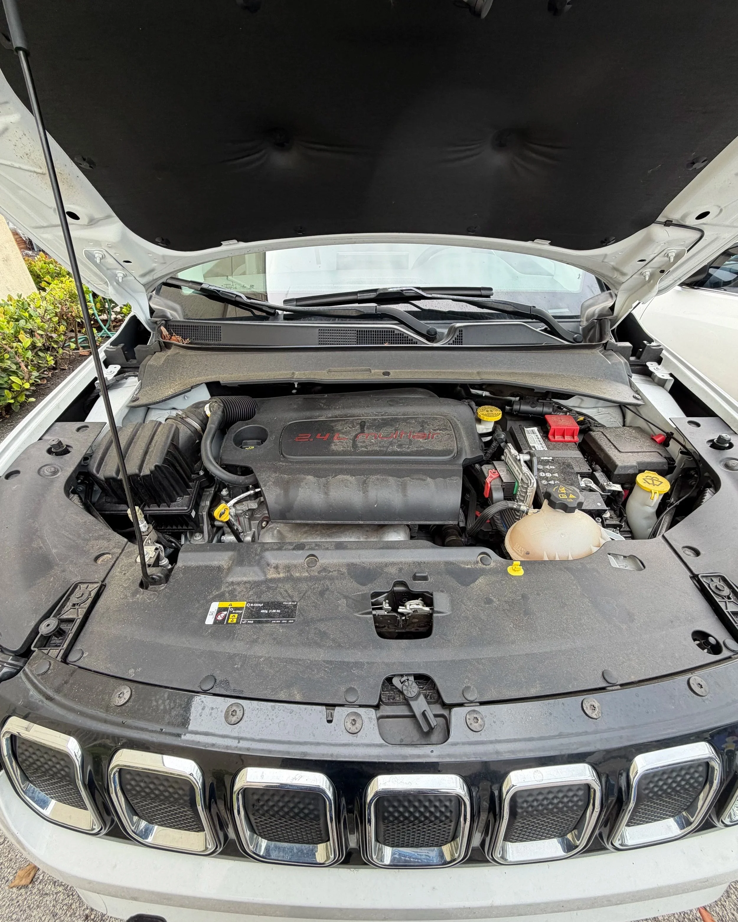Front view of a car with the hood open, revealing the engine bay. The car has a silver exterior, and part of the front grille with vertical slats is visible at the bottom of the image.