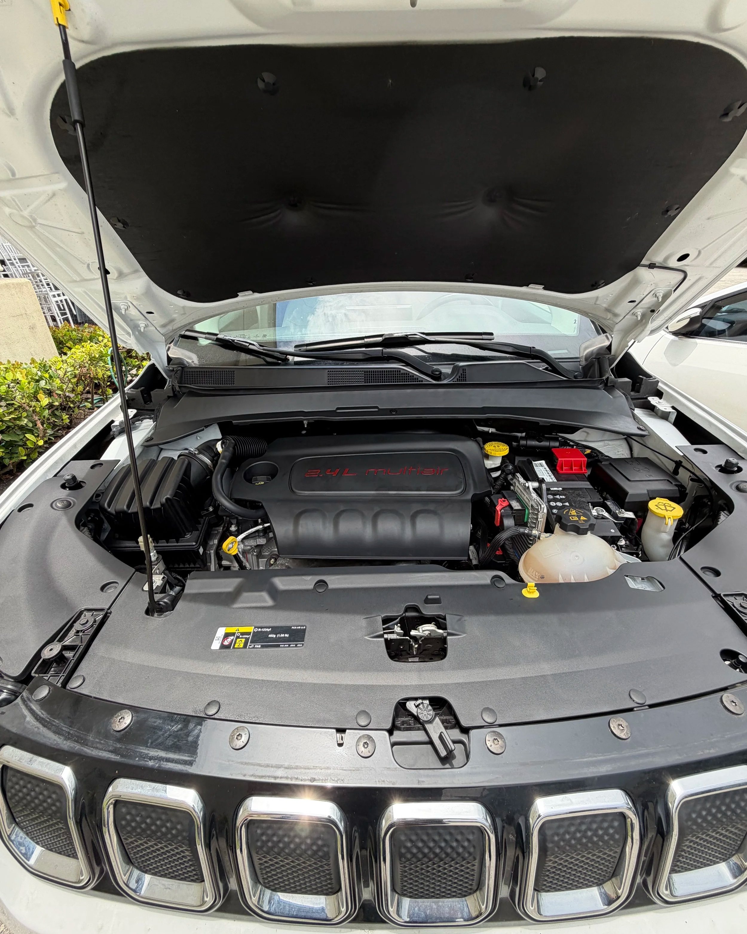View of a car engine under the open hood, with visible components including the air filter, engine cover labeled '2.4L Multiair,' and various fluid reservoirs, with the front grille of the car visible at the bottom.