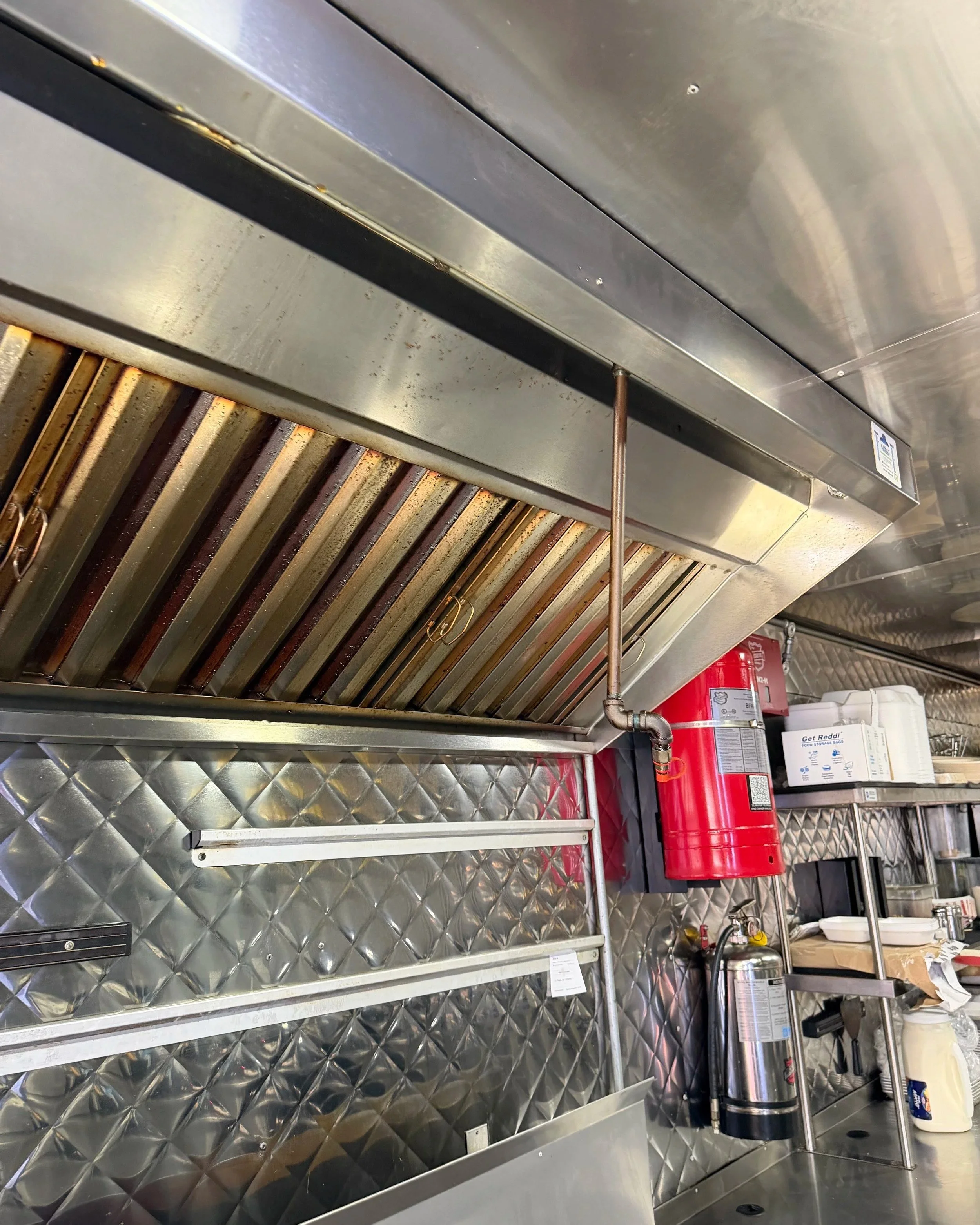 Commercial kitchen with stainless steel ventilation hood and equipment, fire extinguisher, kitchen tools, and supplies.