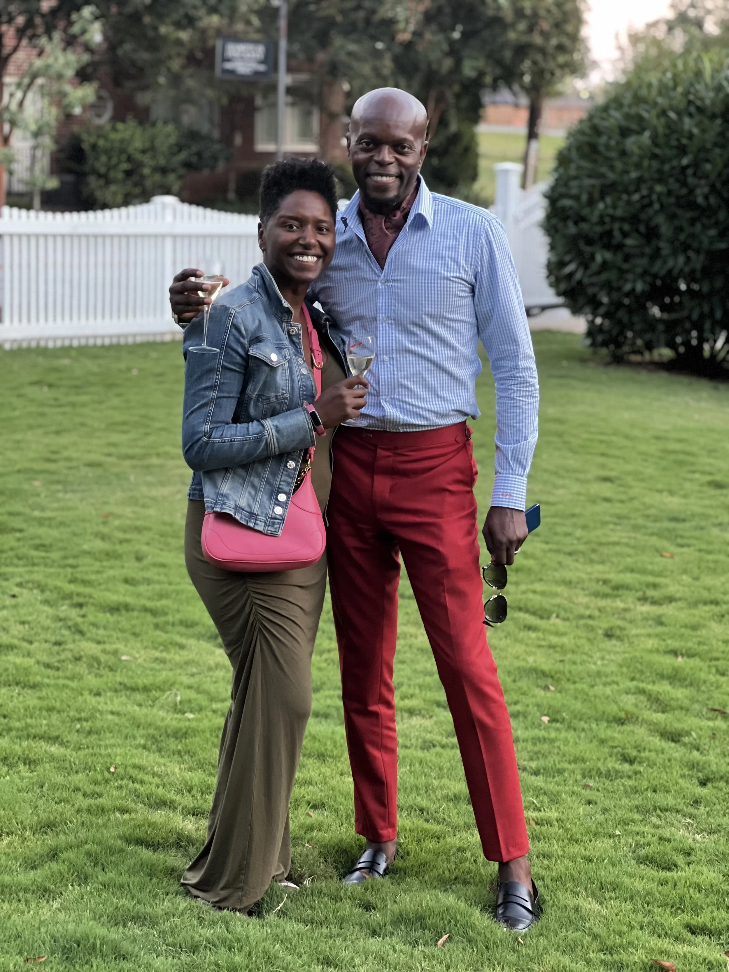 A woman and a man standing on a lawn, smiling at the camera, holding wine glasses, with trees and a white fence in the background.
