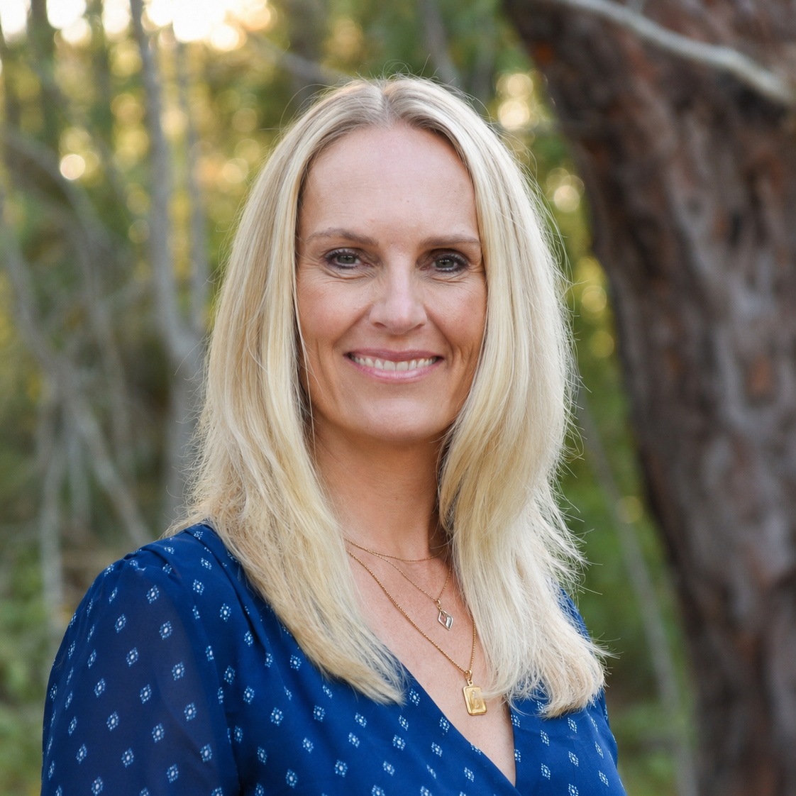 A woman with long blonde hair smiling outdoors with trees and sunlight in the background.