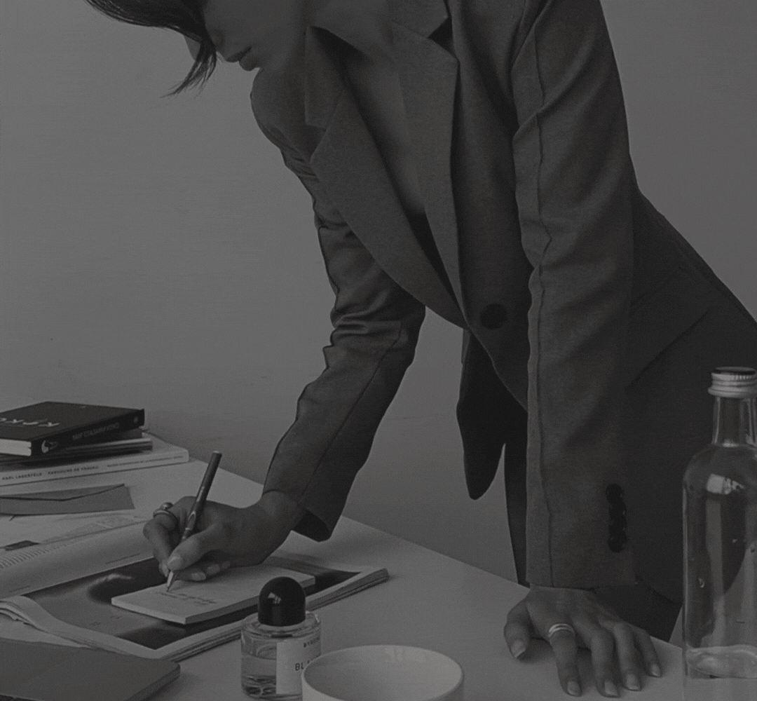 A person in a suit writing on a notepad with various books, a bottle, a small bottle, and a bowl on the table