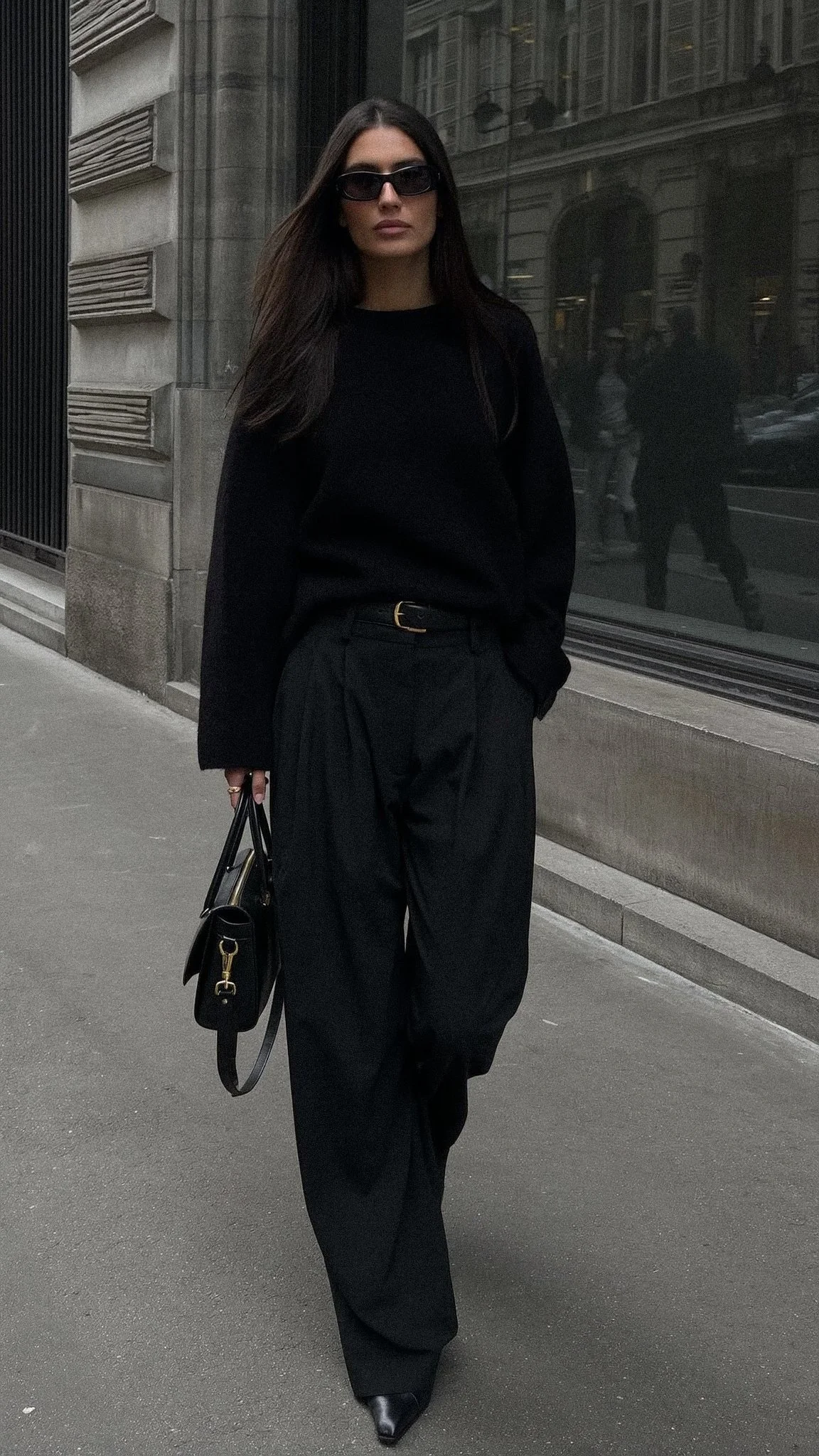 A woman dressed in all black, wearing sunglasses, holding a handbag, walking along an urban street with reflections of buildings and pedestrians in the glass window behind her.