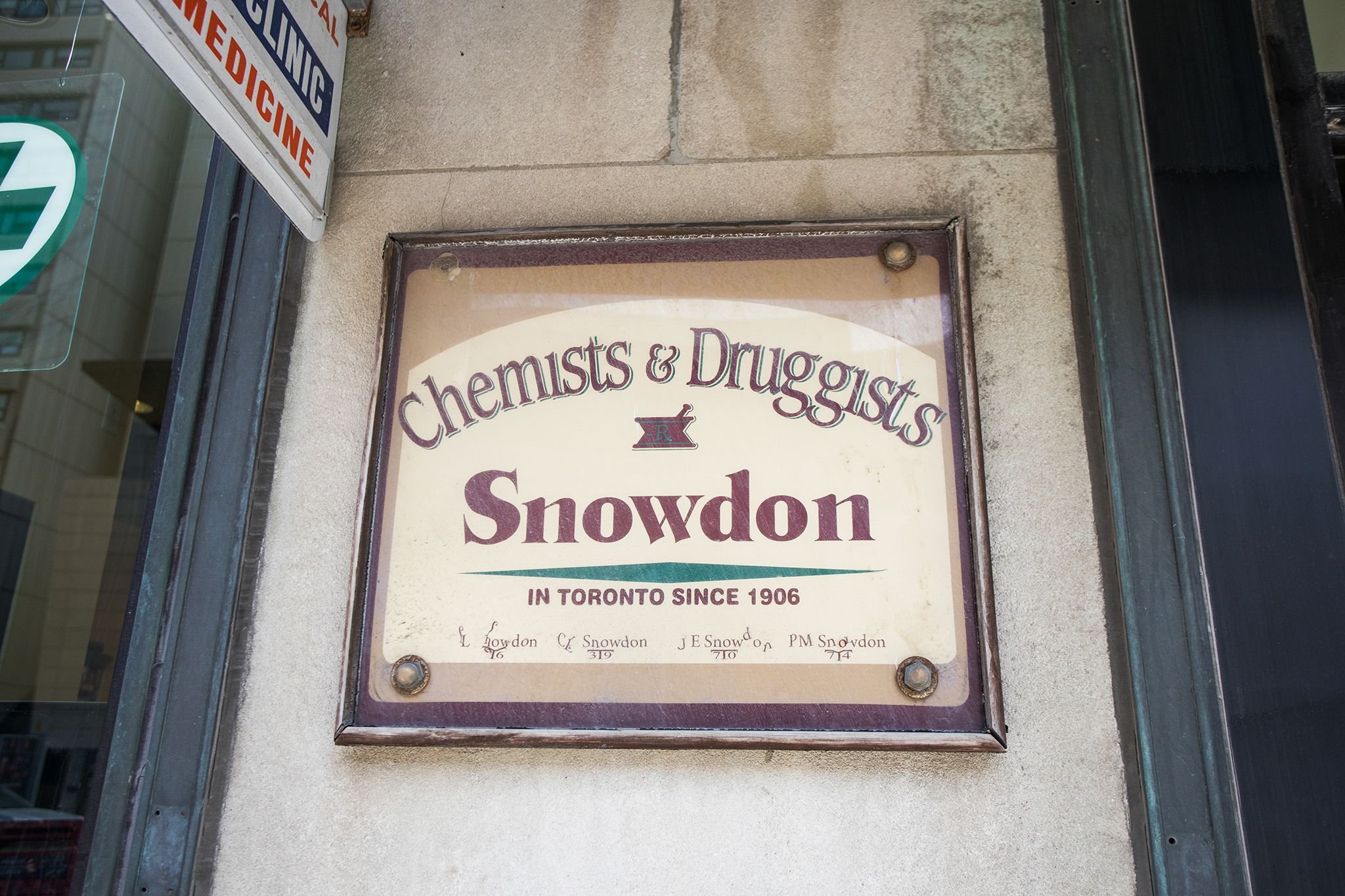 A framed sign on a building's exterior wall advertising 'Chemists & Druggists Snowdon'. It states that the store has been in Toronto since 1906 and includes an illustration of a mortar and pestle.