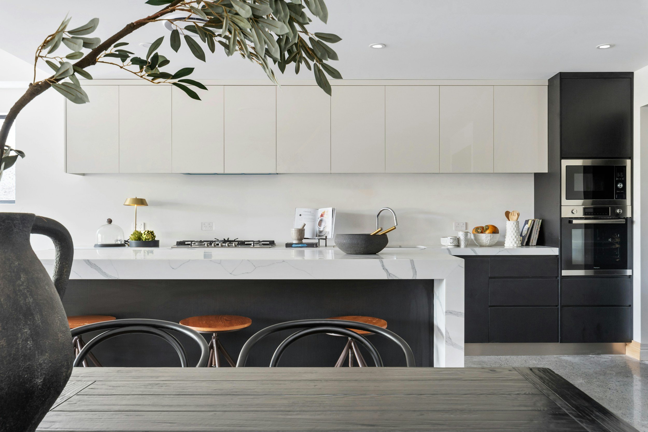 Modern minimalist kitchen with white cabinets, black appliances, marble countertop, and decorative items including a small gold lamp and bowls on the counter, with olive branch plant in the foreground.