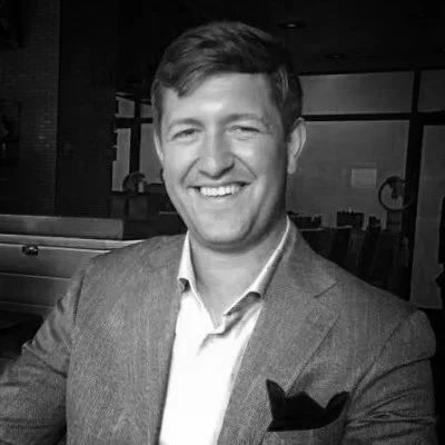 A smiling man in a suit jacket and dress shirt sitting in a modern office environment with large windows.