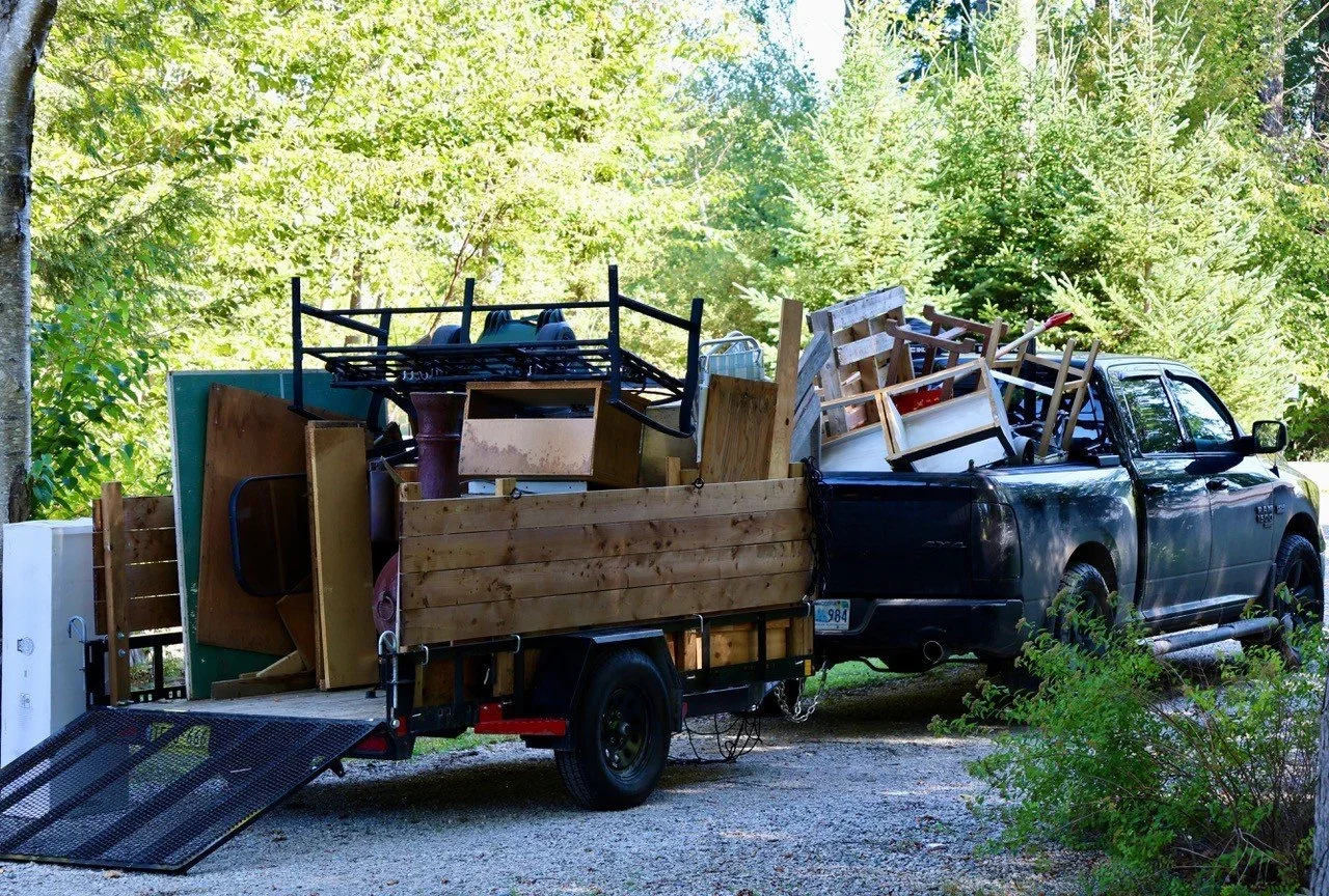 A black truck hauling a trailer filled with furniture and miscellaneous items, including chairs, wooden panels, and a small metal cart, parked on a gravel driveway surrounded by green trees.
