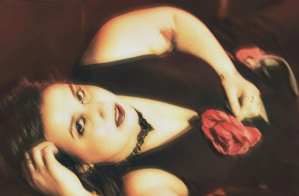 A woman with long dark hair lying on her side on a dark surface, wearing a black top with a red rose on it and a black choker necklace.