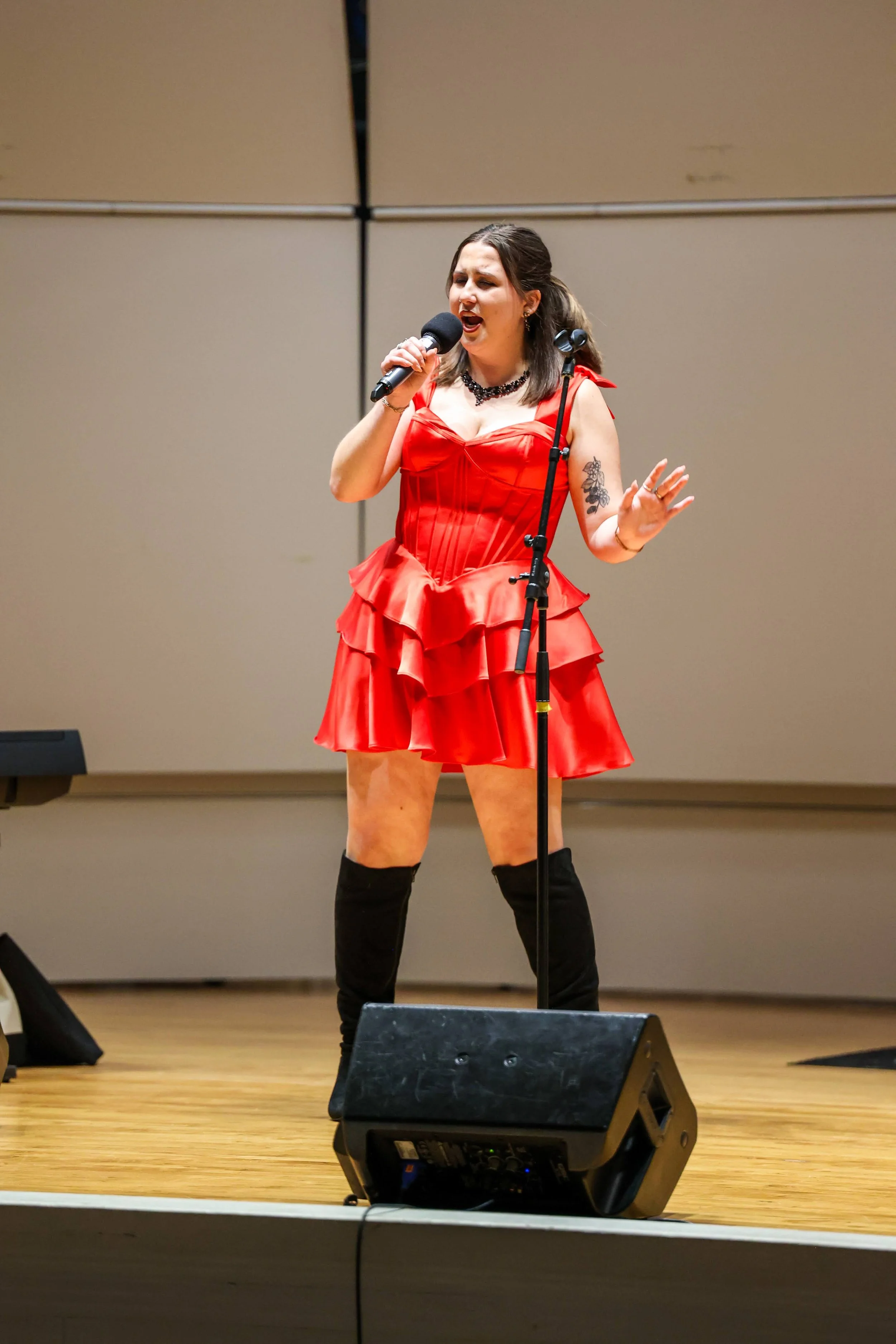 A woman singing on a stage, wearing a red dress, black knee-high boots, and a necklace, with a tattoo on her arm and a microphone in her hand.