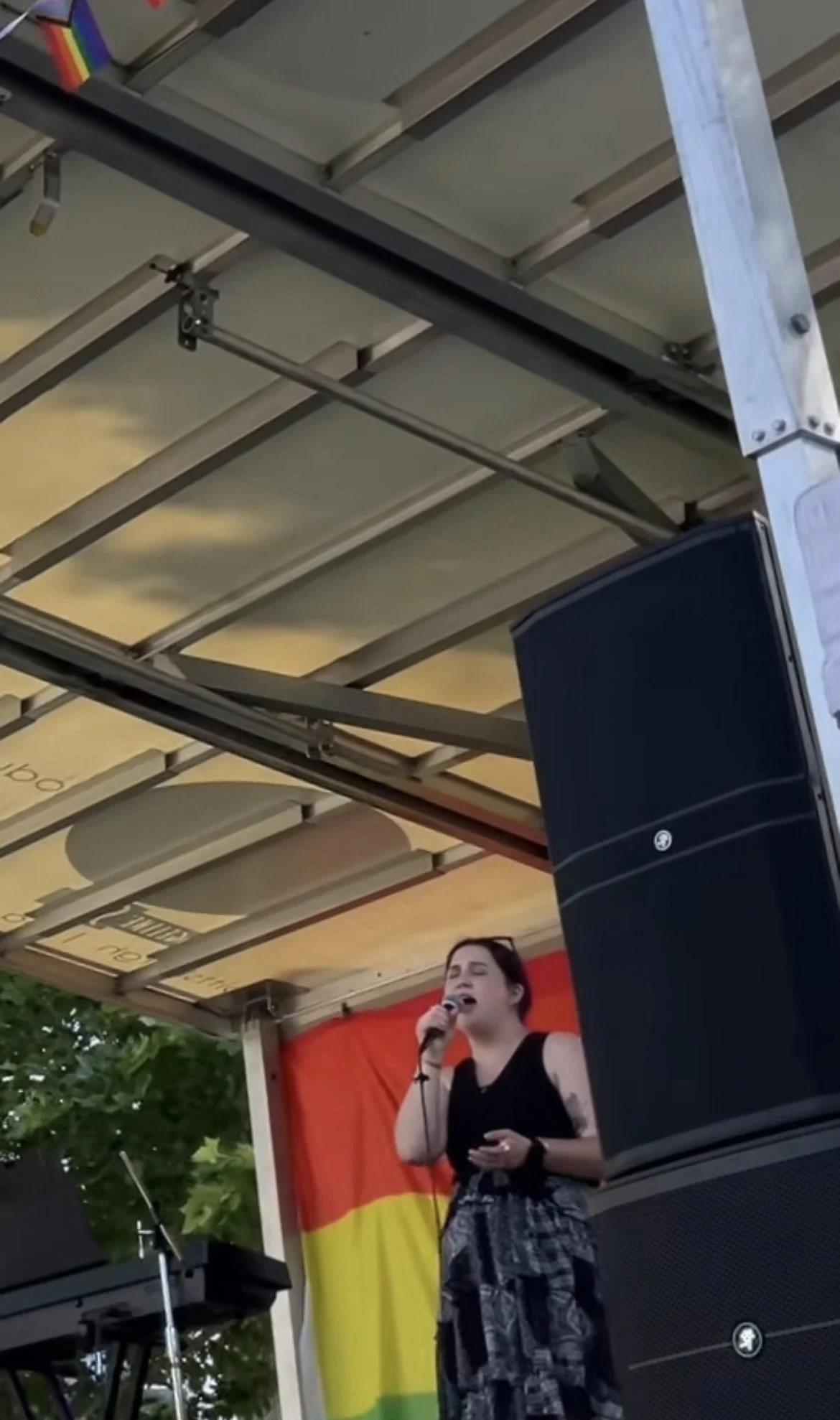 A woman singing into a microphone on an outdoor stage with a colorful backdrop and a rainbow flag. A large black speaker is in front of her, and a keyboard is to her left. The stage has a wooden roof and green trees are visible in the background.