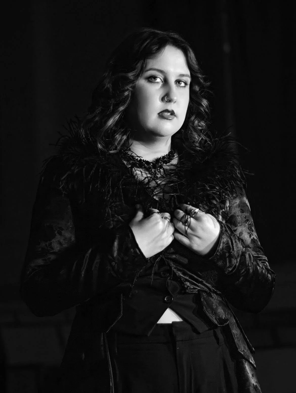 A black and white portrait of a woman with wavy hair, wearing dark makeup, a nose ring, and multiple rings on her fingers, dressed in a textured, feathered garment.