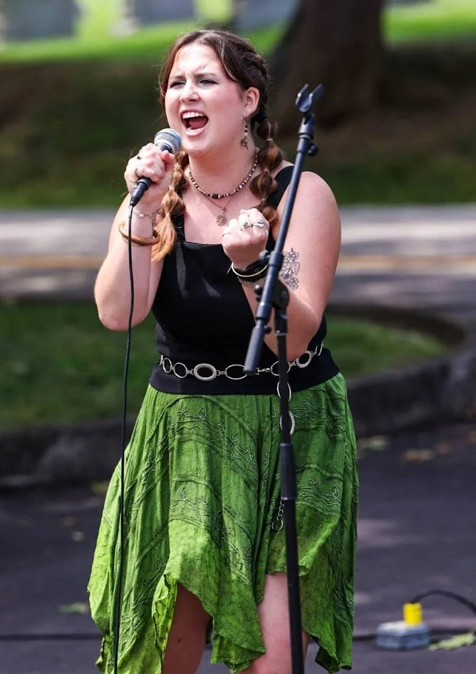Young woman singing passionately into a microphone outdoors, wearing a black top, green skirt, and jewelry, with a tree and grass in the background.