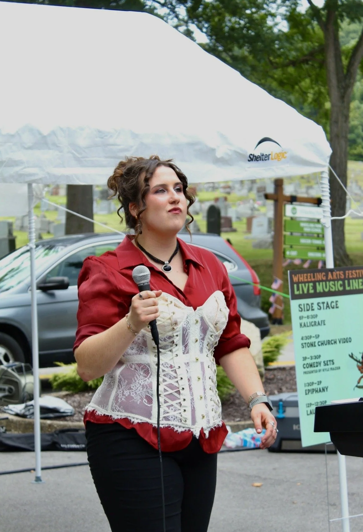 A woman in a red blouse and lace-up corset holding a microphone at an outdoor event, standing in front of a sign with event schedule details at a cemetery.