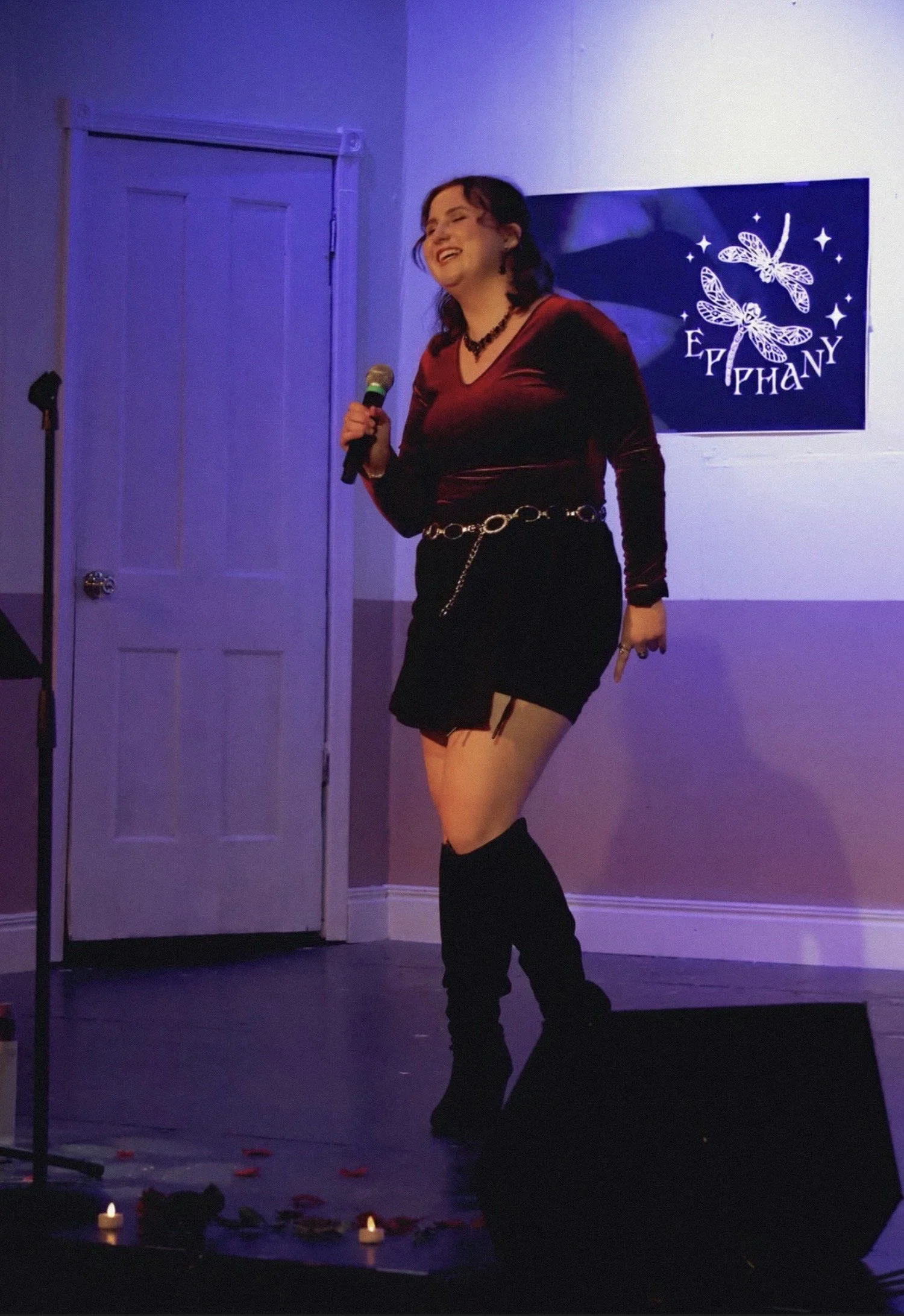 A woman performing on stage, holding a microphone, wearing a red velvet top, black skirt, and thigh-high boots, with a backdrop displaying the name 'Ephany' and dragonfly graphics. Candles and rose petals are arranged on the floor in front of the sta