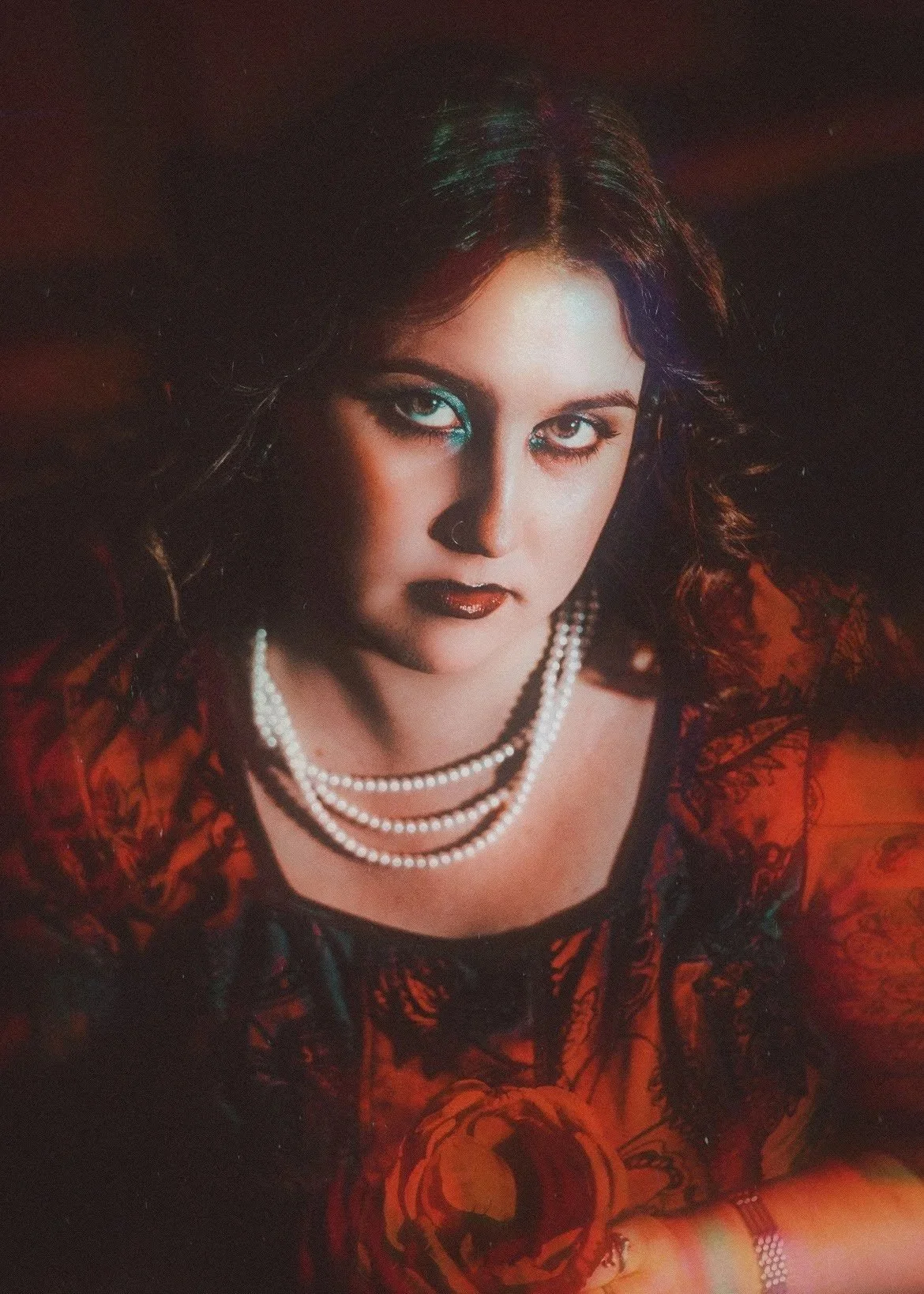 A woman with dark hair and piercing eyes wearing a red and black patterned dress, layered pearl necklaces, and jewelry, with intense makeup and a dark background.
