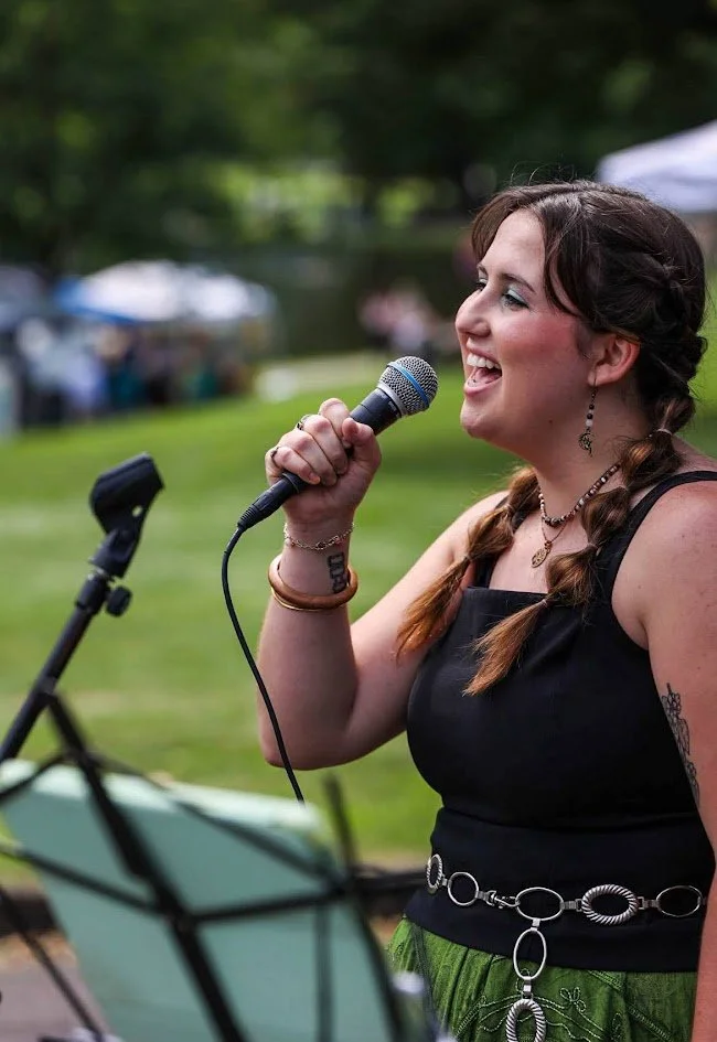 A woman singing into a microphone outdoors at a park or festival setting, with a music stand in front of her.