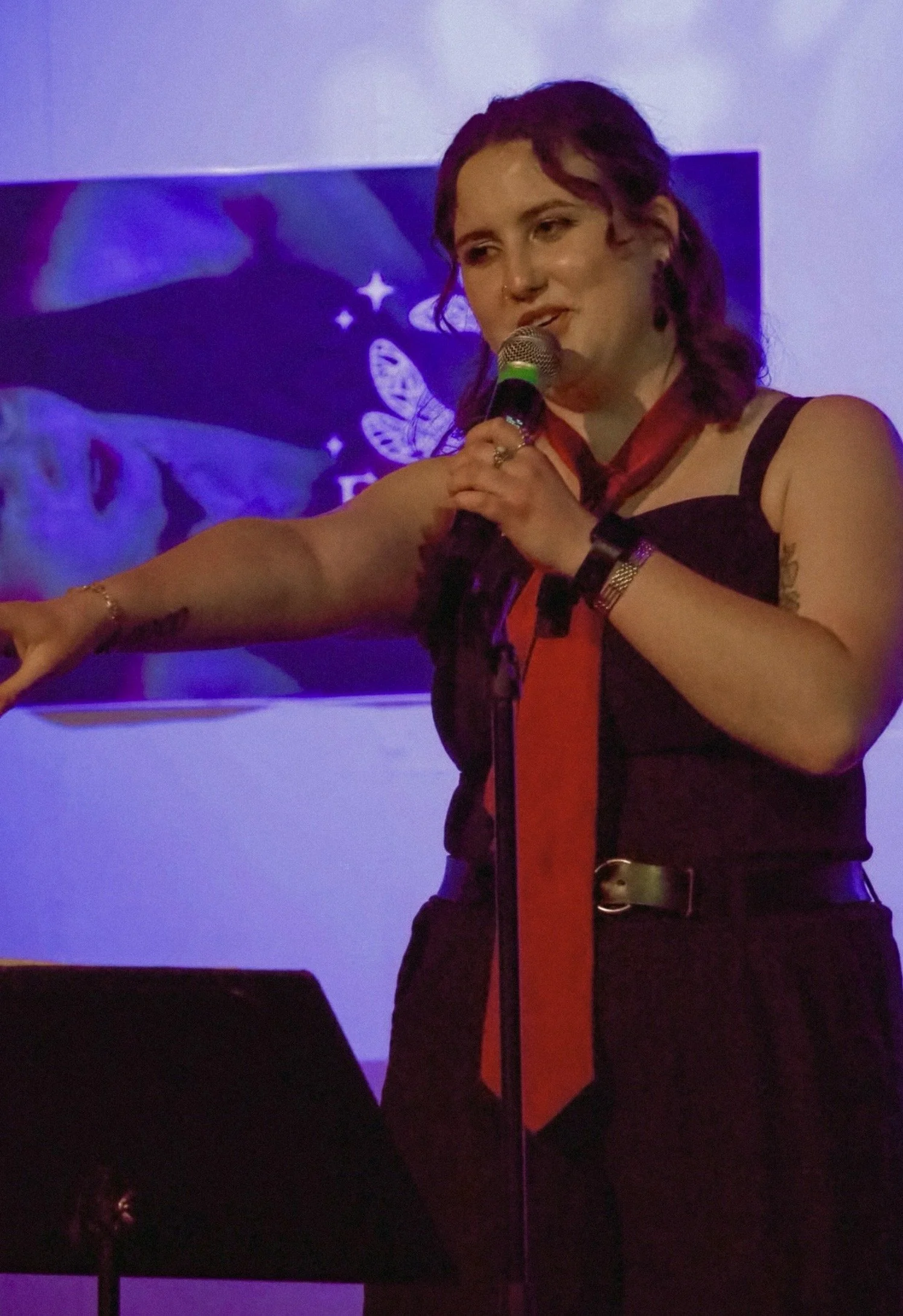 A woman with dark hair and tattoos on her arm is speaking into a microphone during a presentation or event, with blue and purple lighting and a blue banner with a butterfly or insect design in the background.