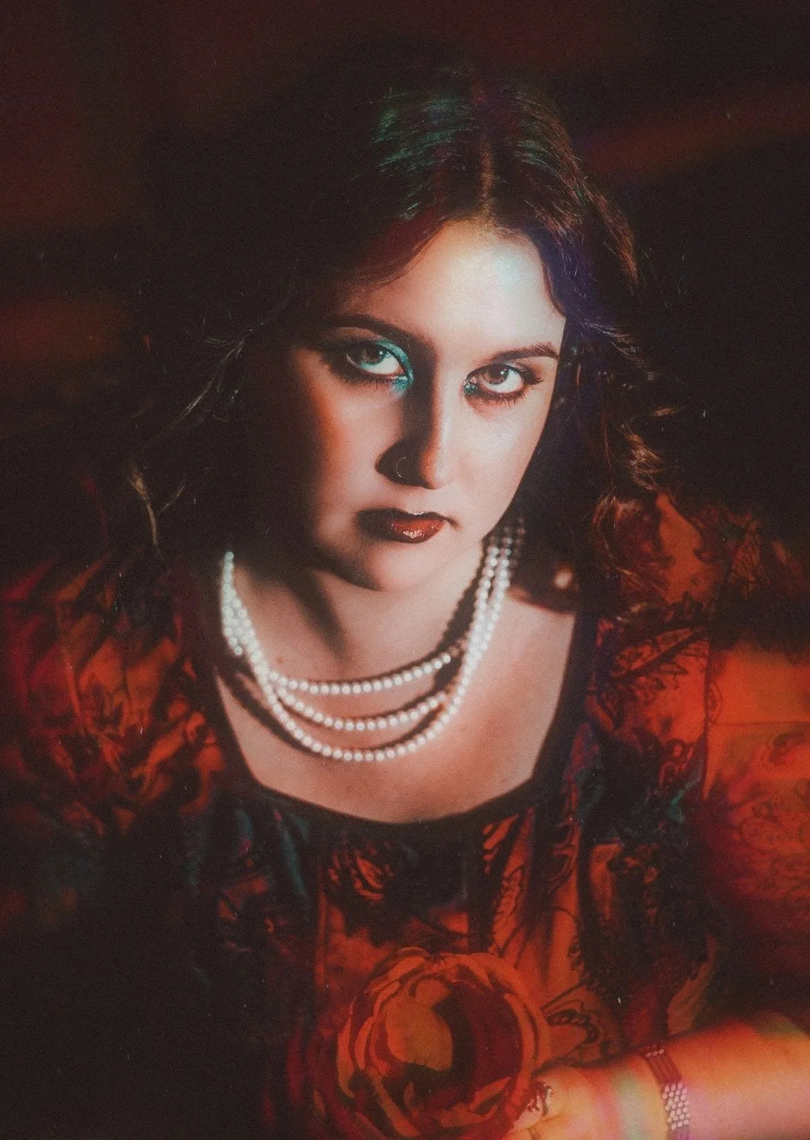 A woman with dark hair and intense eye makeup wearing a red patterned outfit and a string of pearls.
