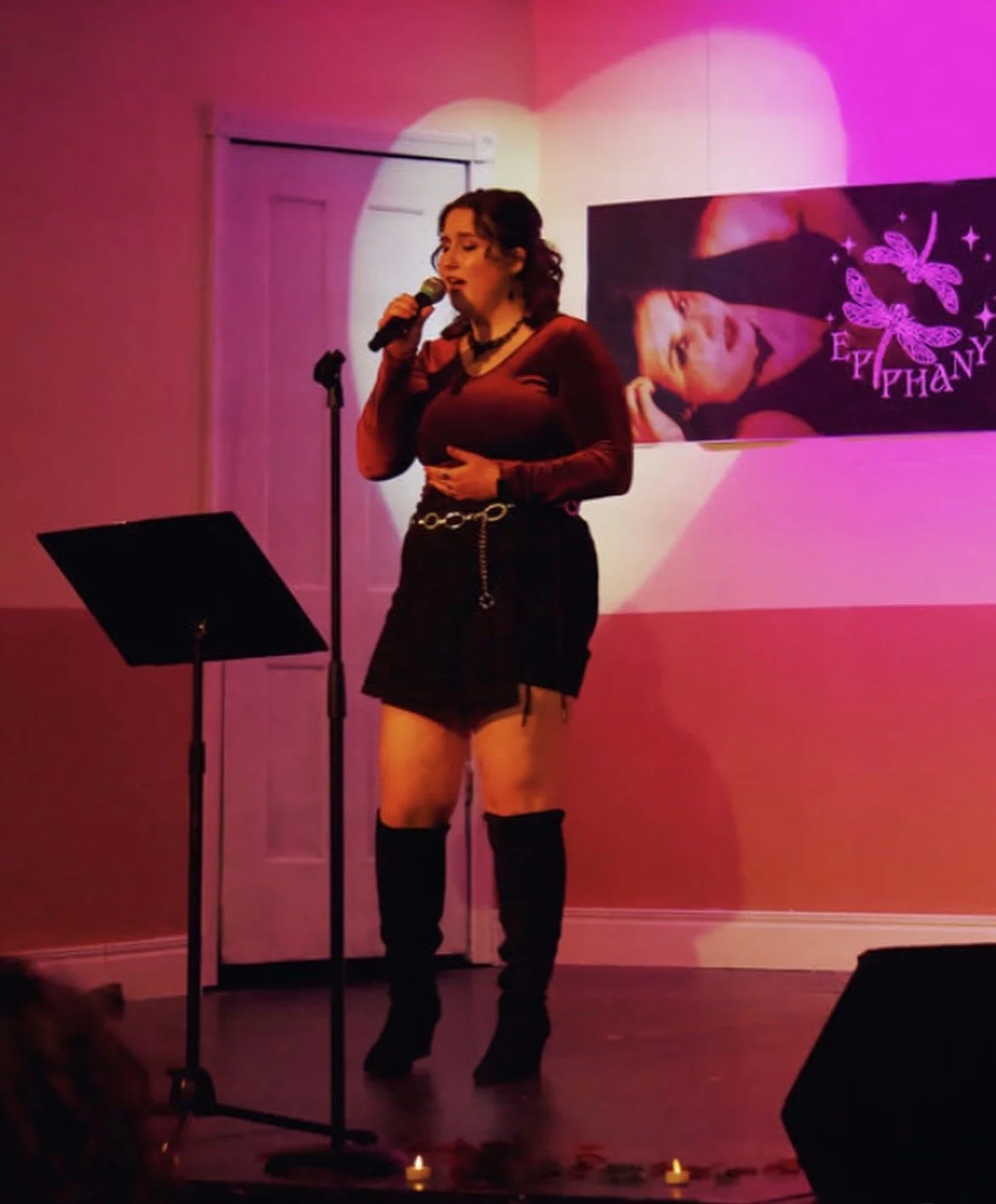 A woman singing into a microphone on a stage with pink and purple lighting. Behind her, a large poster of herself with the name 'Ephany' and butterfly graphics is displayed.