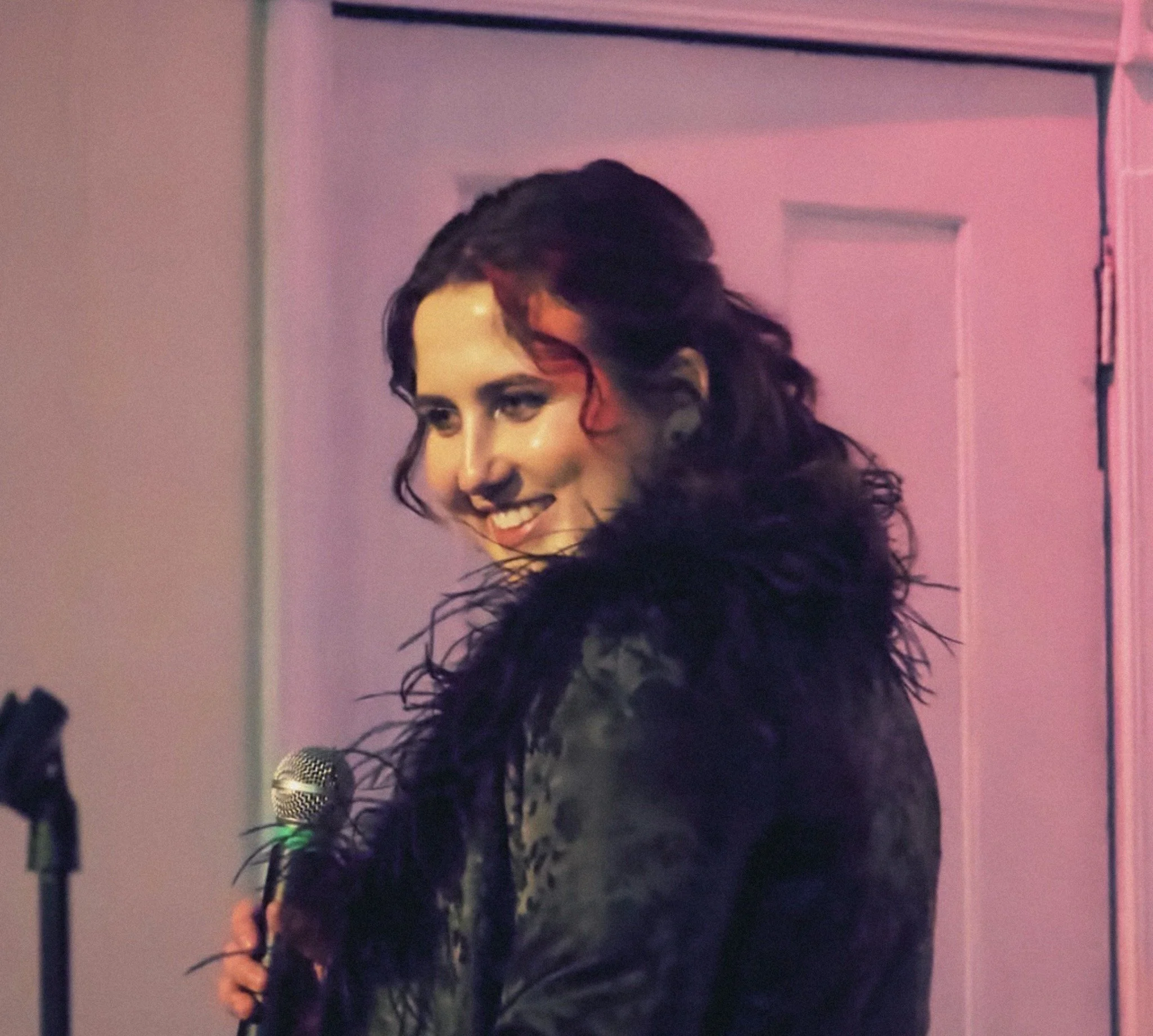 A woman with curly dark hair smiling, holding a microphone, standing in front of a pink and purple background.