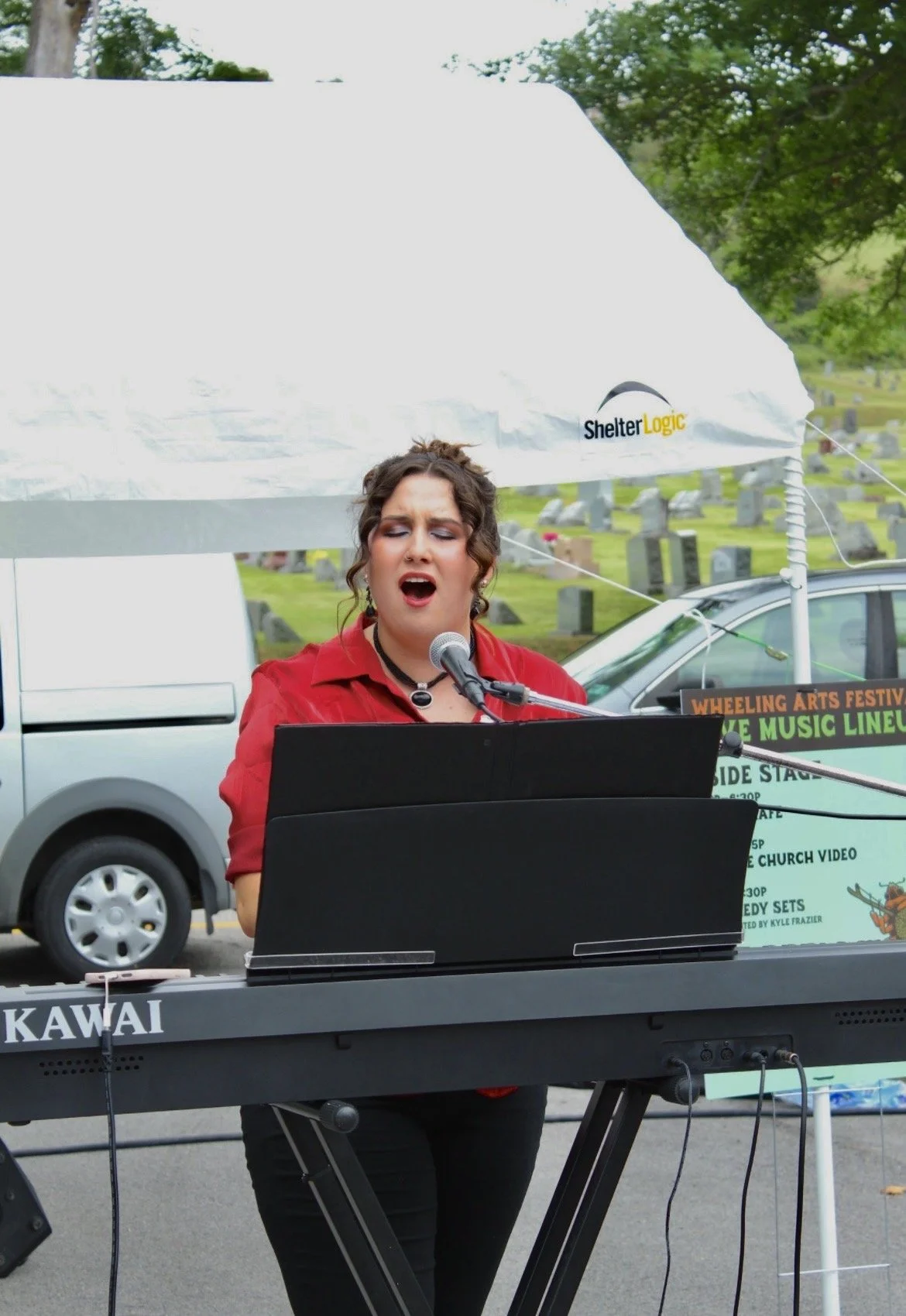 A woman singing into a microphone while playing a keyboard outdoors at an event. She has dark curly hair and is wearing a red top and black pants. Behind her, there is a white canopy tent with 'Shelter Logic' written on it, a sign with event informat