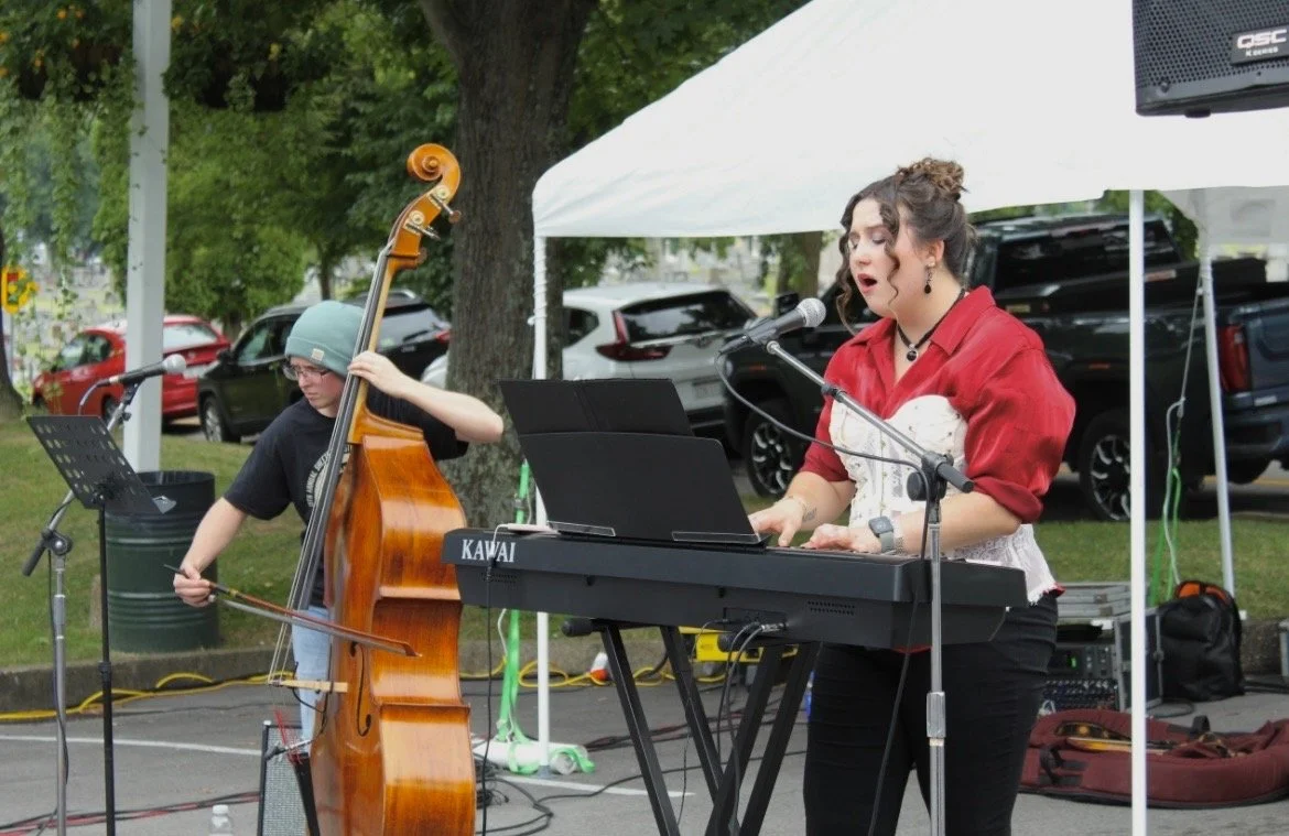 Female musician playing keyboard and singing into a microphone, with a young boy playing double bass in the background, outdoors on a stage with a white canopy.