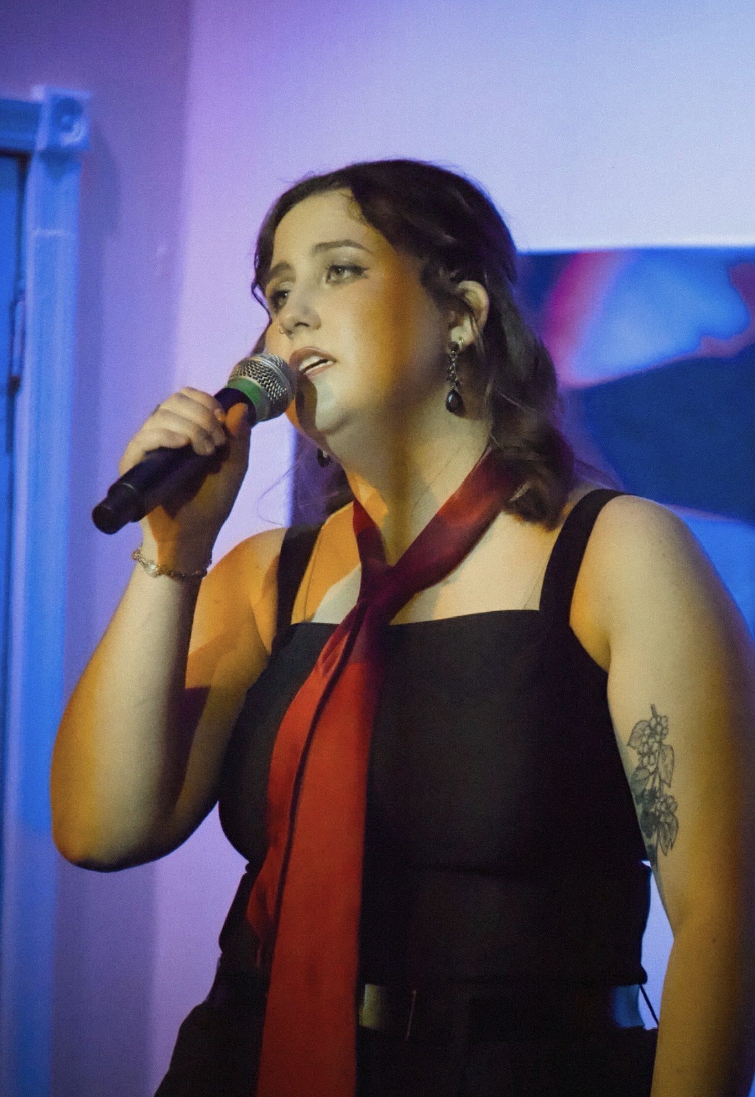 A young woman with dark hair, earrings, and a tattoo on her arm, is singing into a microphone with a colorful backdrop.