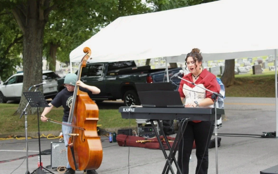 A woman singing and playing a keyboard outdoors with a young boy playing a double bass beside her. They are performing at an event with a white canopy tent and parked cars in the background.