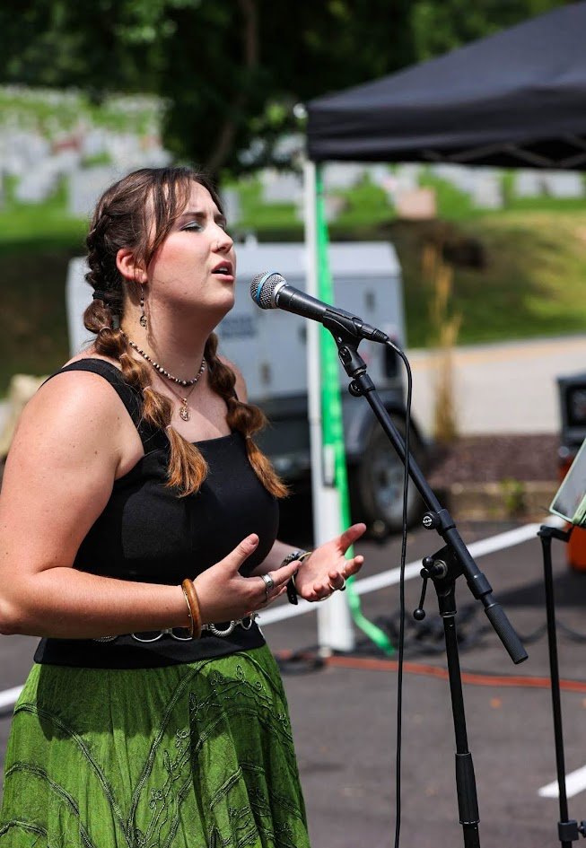 A young woman with braided hair singing into a microphone outdoors during daytime, with greenery, a tent, and a vehicle in the background.