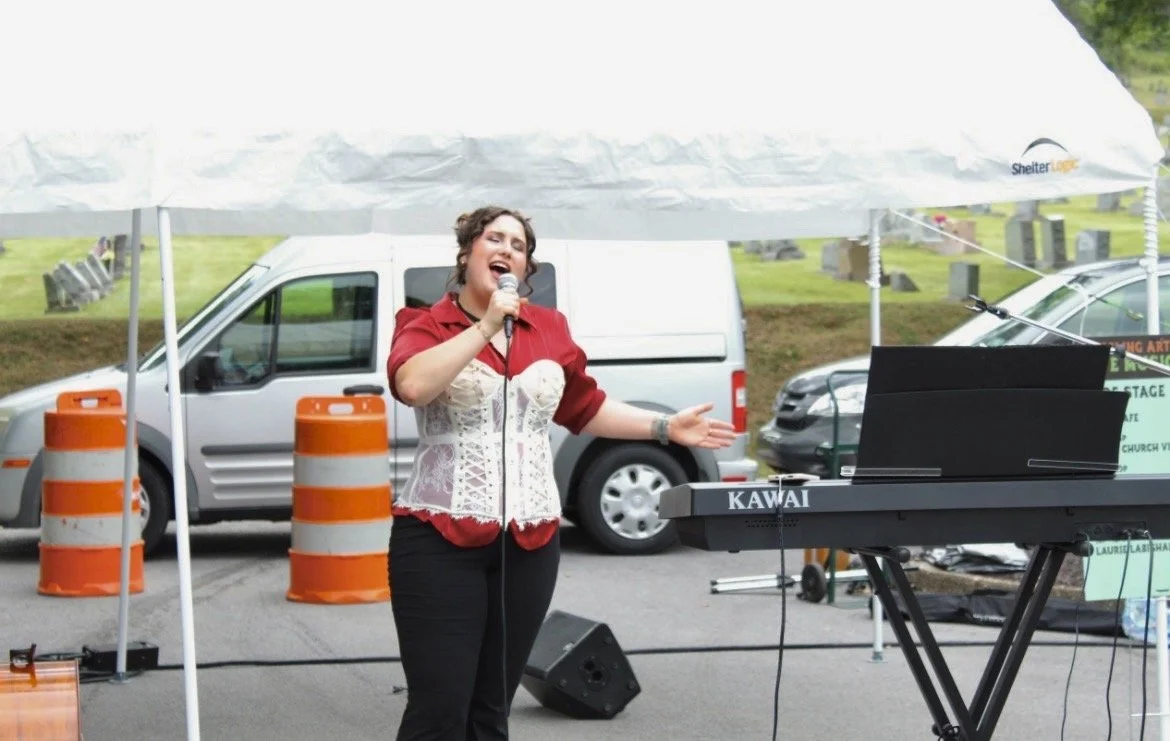 A woman singing into a microphone while standing next to a keyboard at an outdoor event, with a white van and cars parked in the background and orange traffic cones nearby.