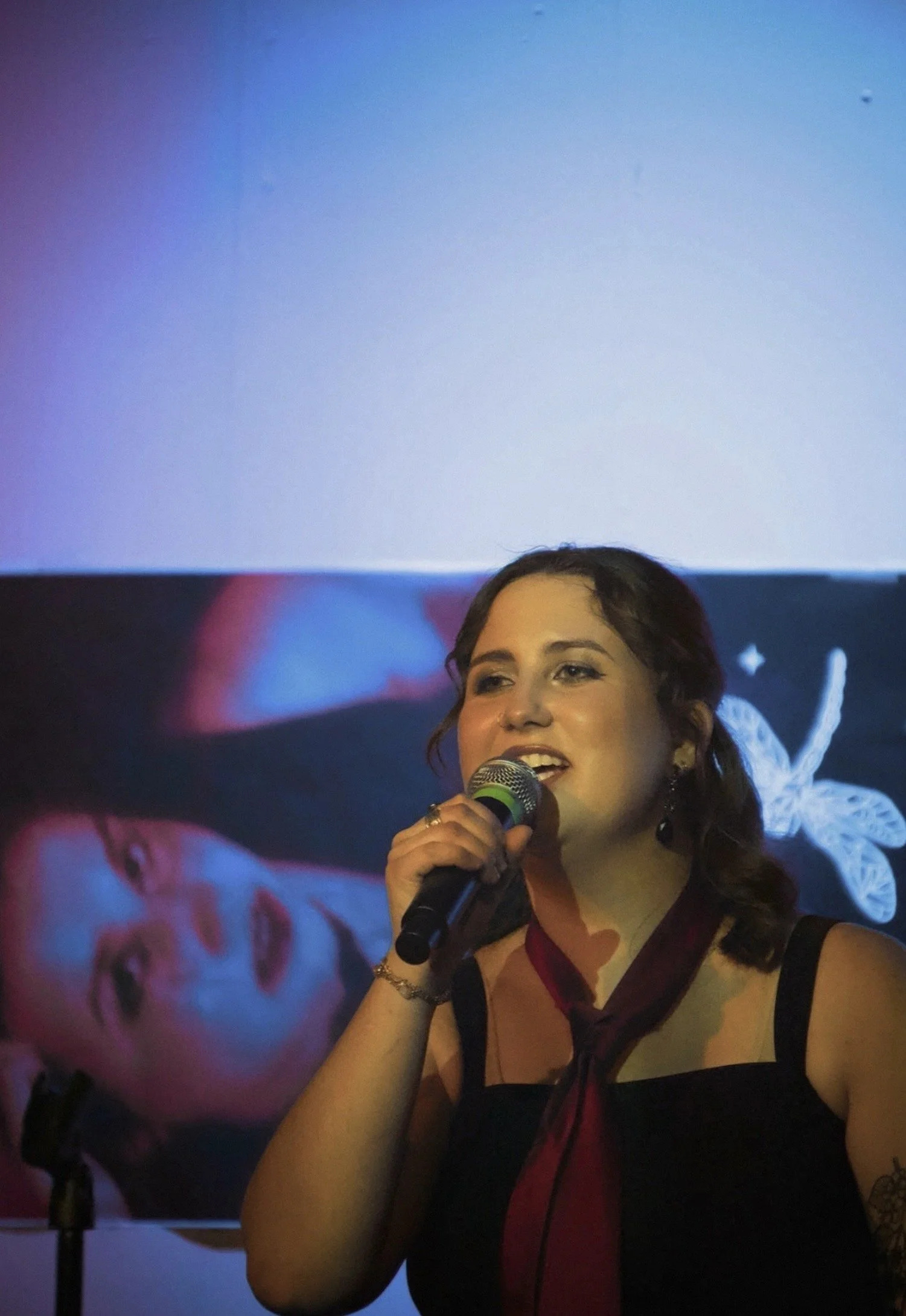 Woman speaking into a microphone at an event with a large digital screen in the background.