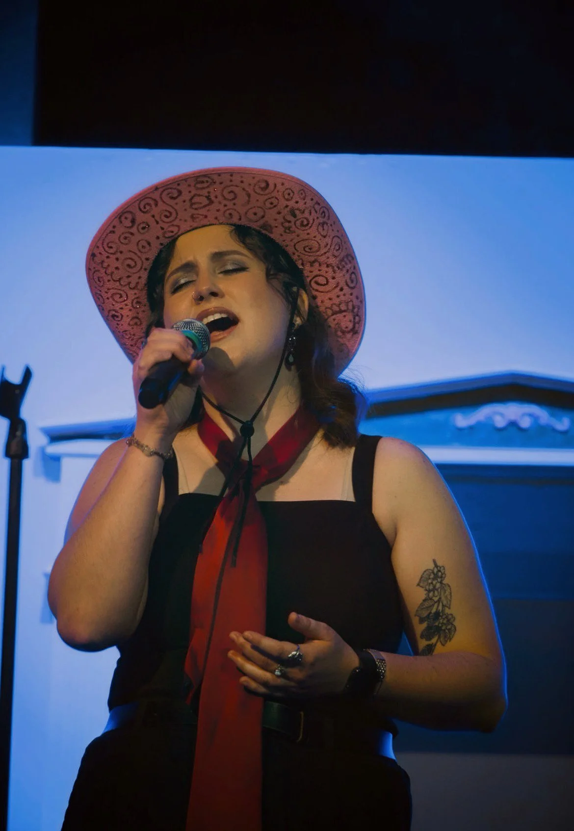 A woman singing on stage, wearing a pink hat with black swirls, a black dress, and a red scarf around her neck. She has a tattoo on her left arm and is holding a microphone with her eyes closed.